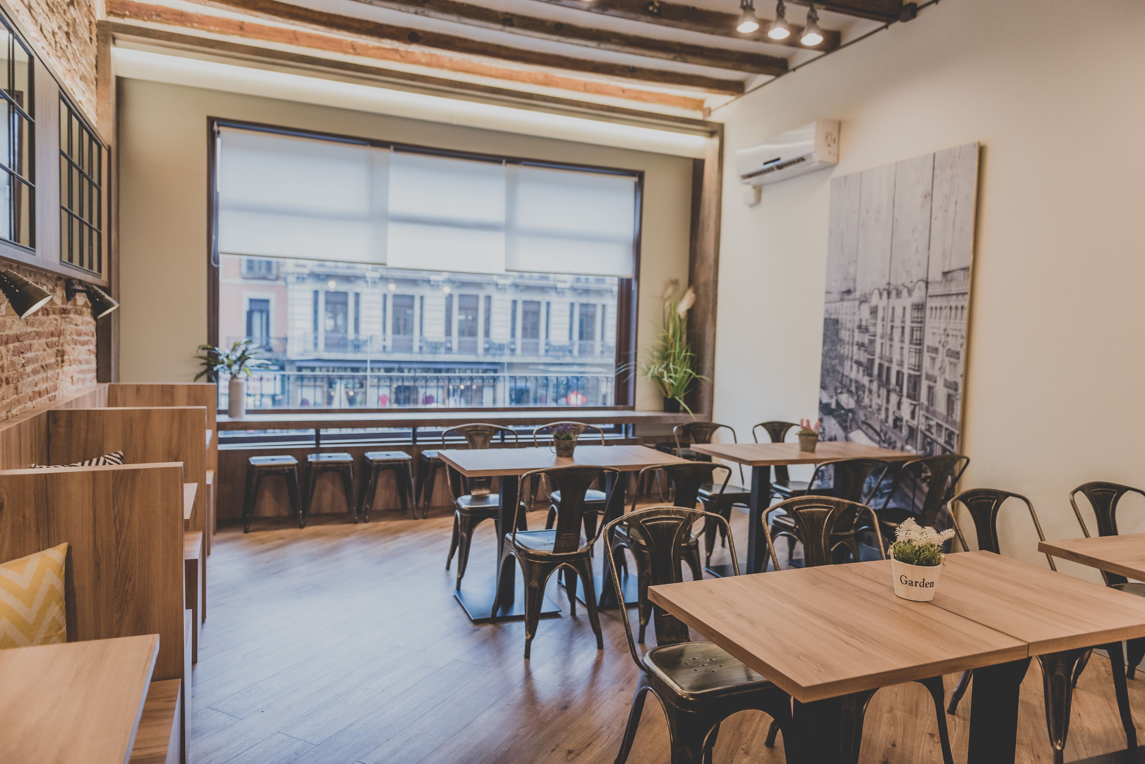 Un restaurant moderne avec des tables et des chaises en bois, une grande fenêtre donnant sur un paysage urbain et un mur de briques sur l’un des côtés.
