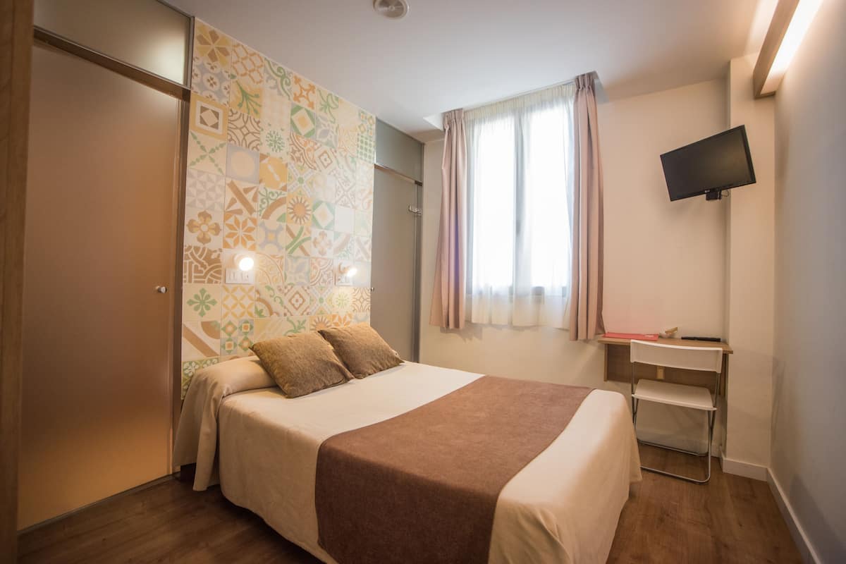 Habitación de hotel con una cama, televisión montada en la pared, escritorio con silla y ventana con cortinas.
