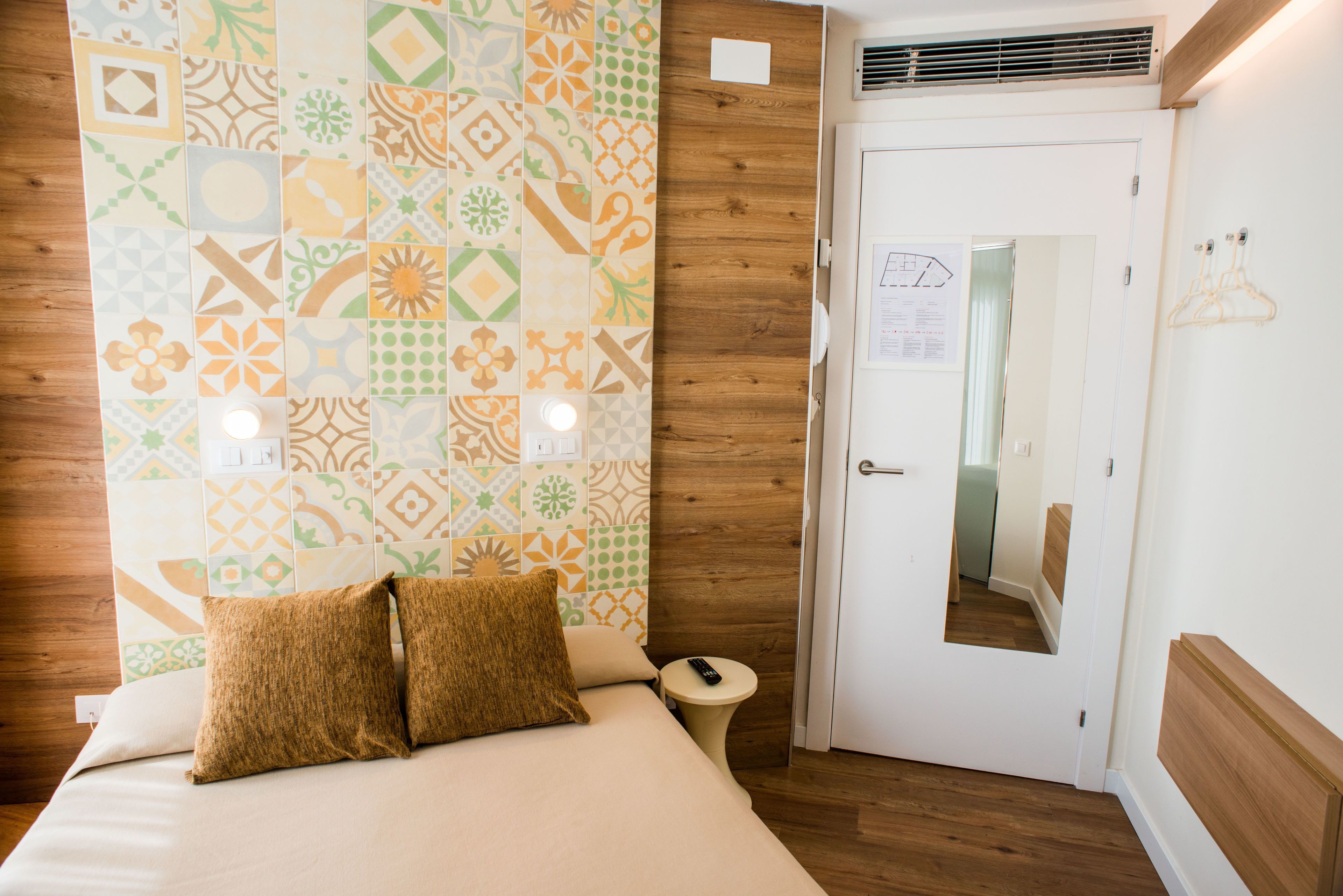 Quarto de hotel com parede estampada, cabeceira de madeira, cama com duas almofadas, mesa de cabeceira com controle remoto e porta com espelho.