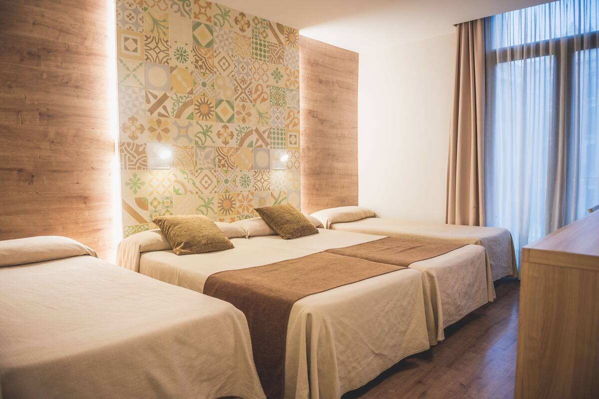 Una habitación de hotel con tres camas, un papel tapiz estampado y una ventana con cortinas.