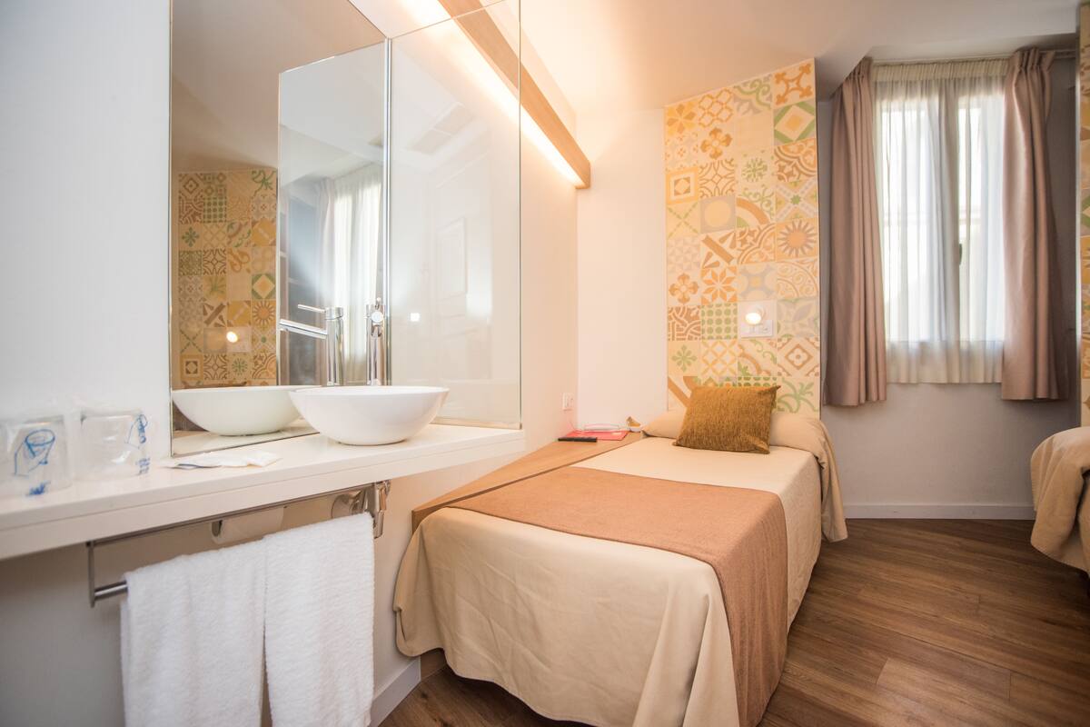 Habitación de hotel con un espejo grande, dos camas, un baño con lavabo y lavatrastos, y papel tapiz estampado.