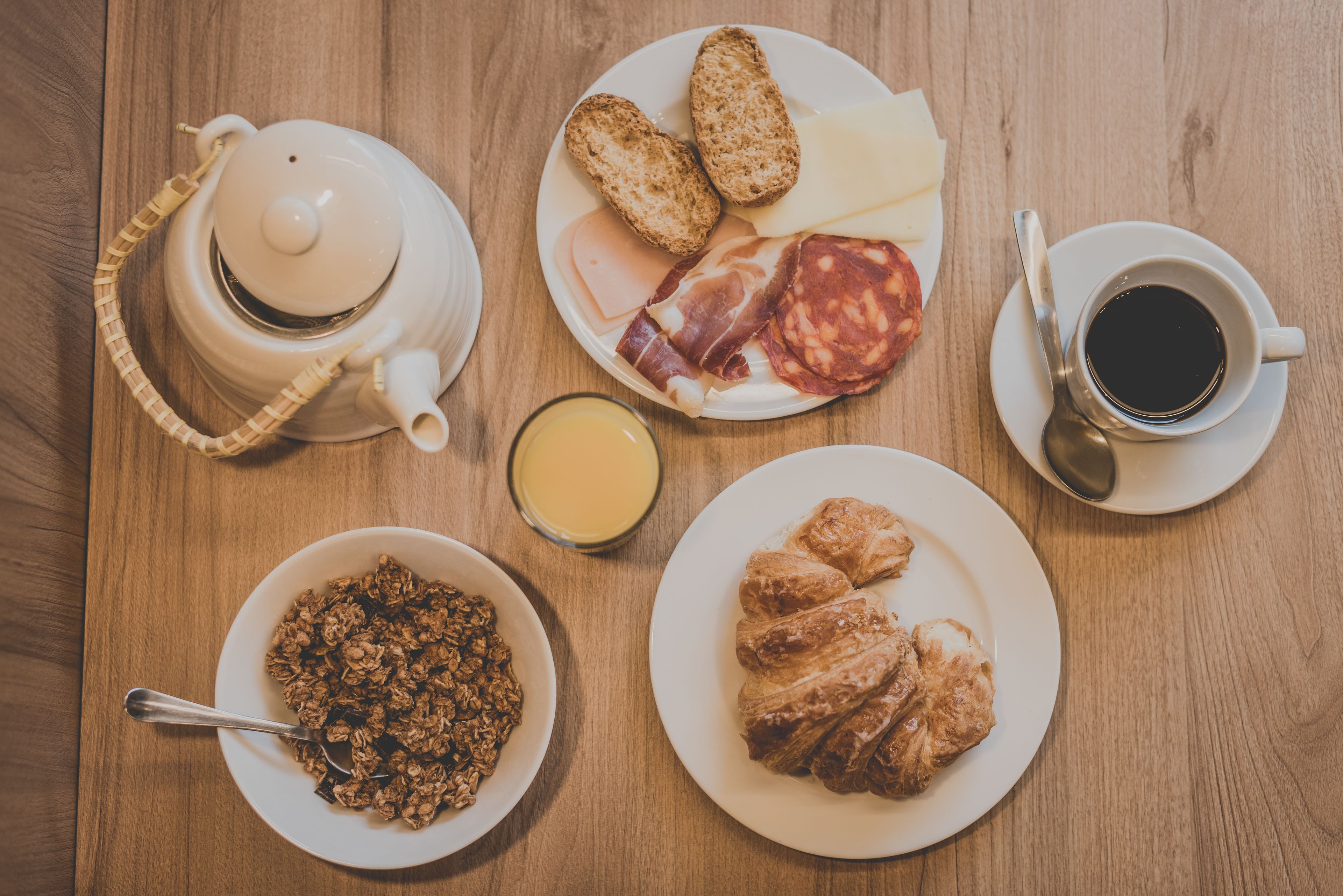 Uma mesa de café da manhã com uma chaleira, torradas, queijo, salame, croissants, uma tigela de cereal, uma xícara de café e um copo de suco de laranja.