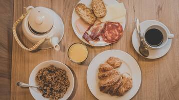 Uma mesa de café da manhã com uma chaleira, torradas, queijo, salame, croissants, uma tigela de cereal, uma xícara de café e um copo de suco de laranja.