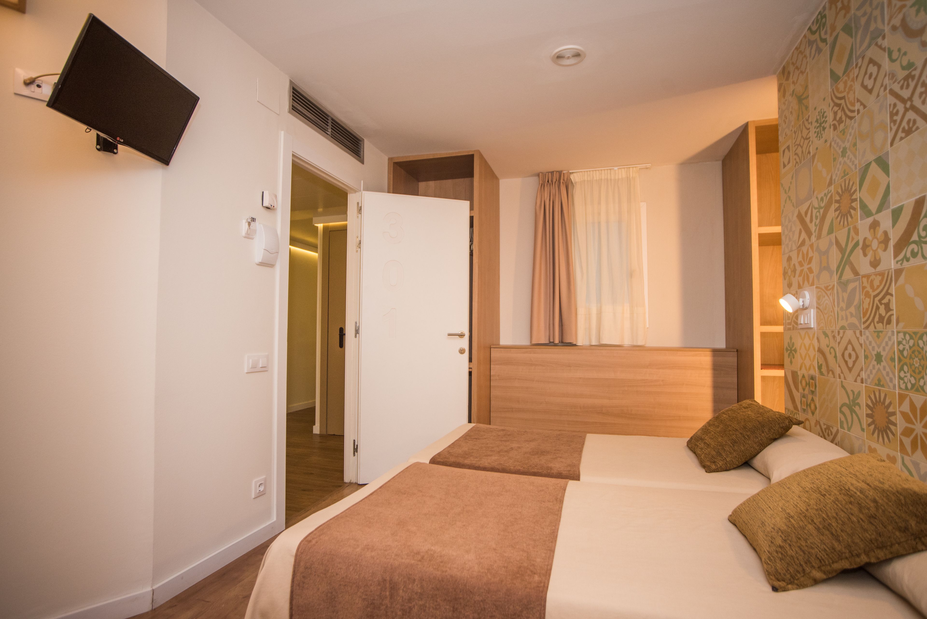A hotel room with two beds, a TV mounted on the wall, a door leading to a bathroom, and patterned wallpaper.