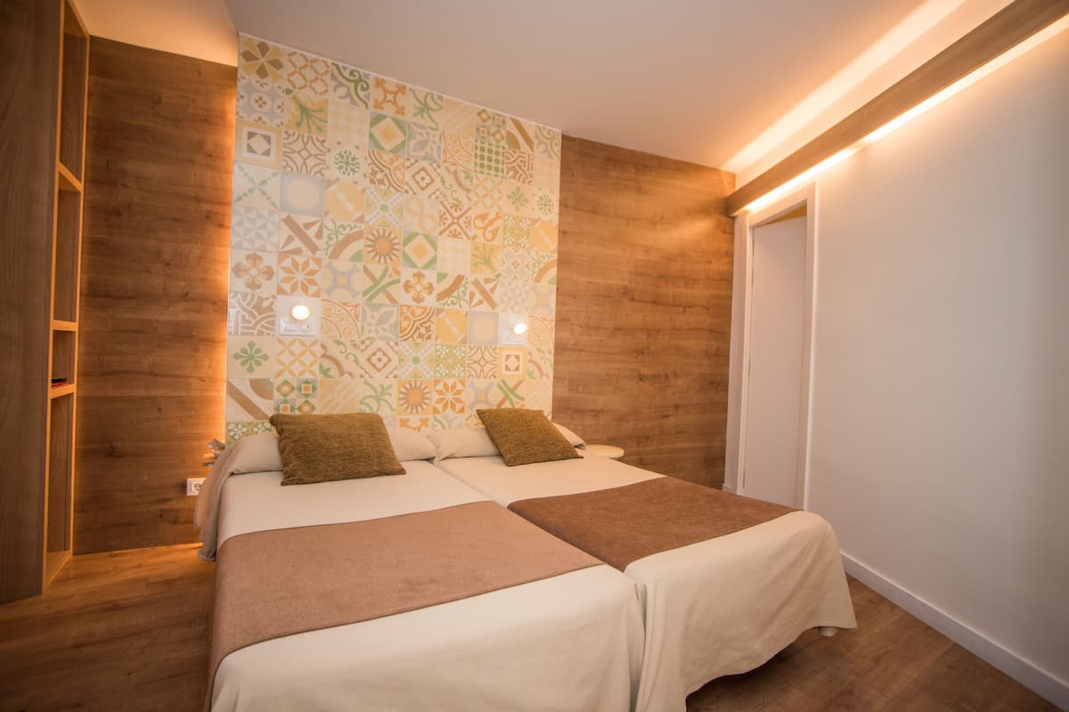 Habitación de hotel con piso de madera, dos camas con sábanas blancas y mantas marrones, una pared estampada y una puerta con interruptor de luz.