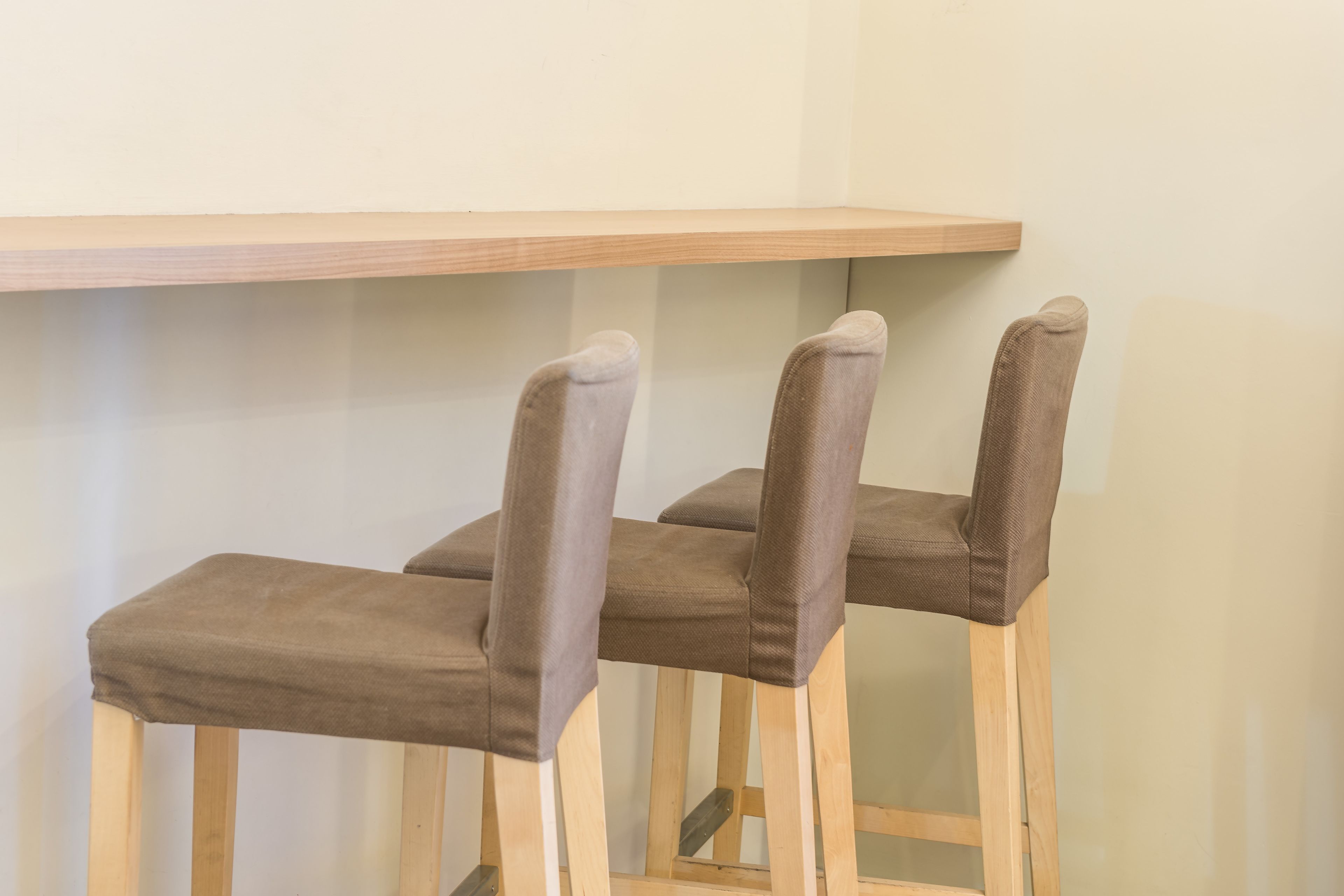 Three wooden chairs with cushioned seats and backs, positioned under a wooden shelf against a plain wall.