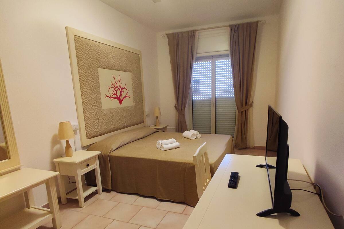 Une chambre d’hôtel comprenant un lit, un bureau, une chaise, une télévision, une lampe, une fenêtre avec des rideaux et un ventilateur de plafond.