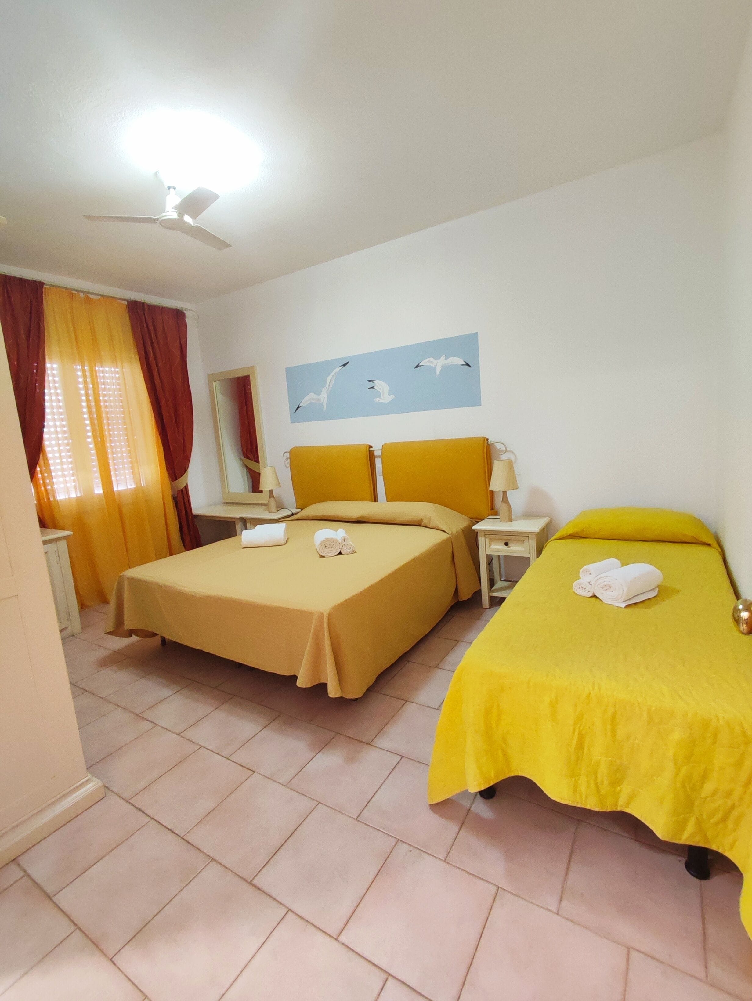 A hotel room with two beds, a ceiling fan, a window with curtains, a wall-mounted mirror, and a painting of seagulls.