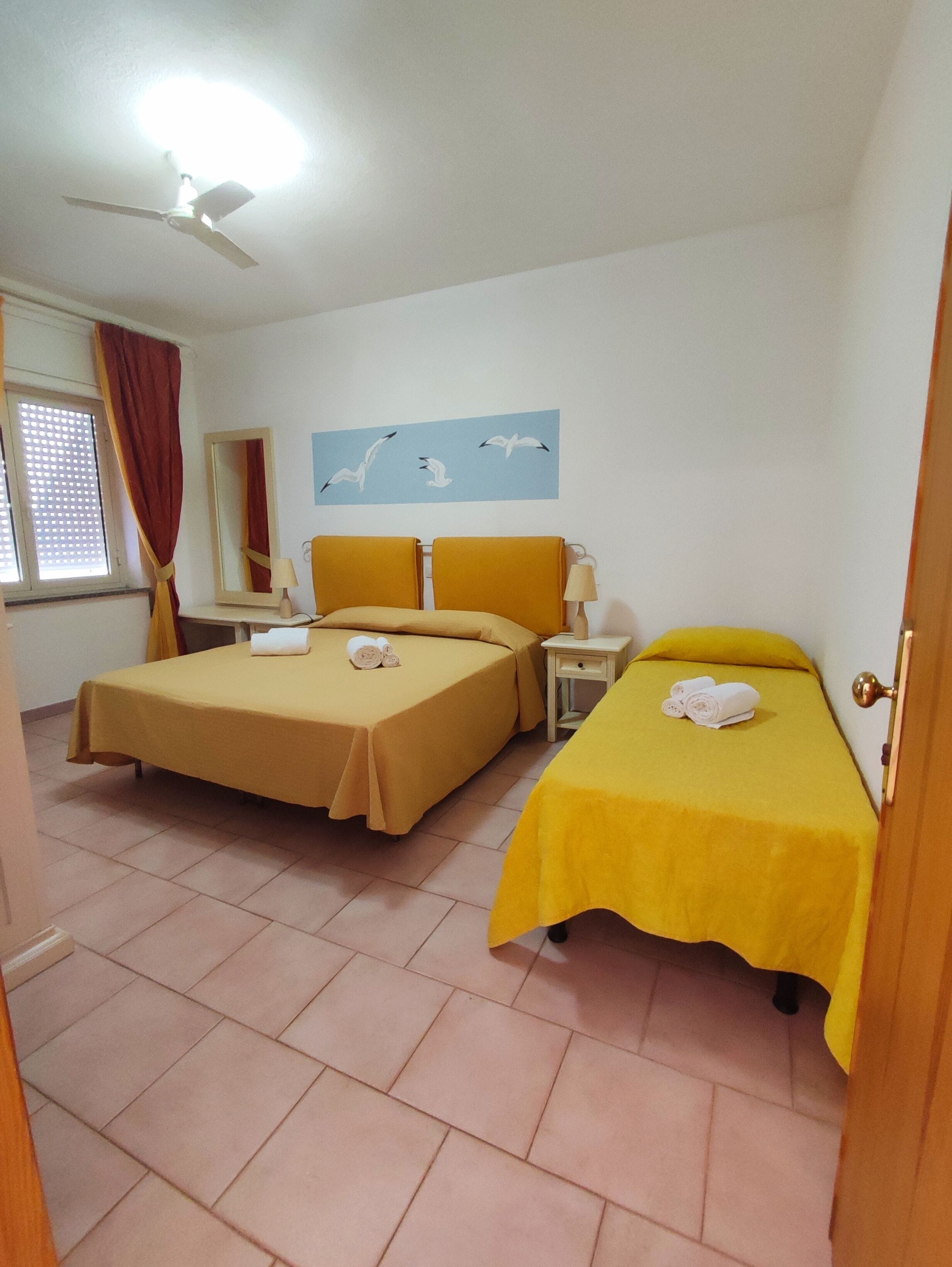 Quarto de hotel com duas camas, um ventilador de teto, janela com cortinas, um espelho e um quadro na parede com gaivotas.