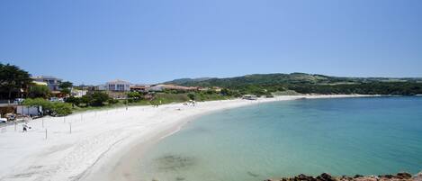 Una playa costera con aguas cristalinas, arena dorada y edificios a lo largo de la costa.