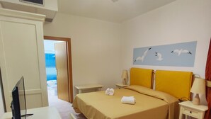 A hotel room with a bed, a desk, a computer, a ceiling fan, and a view of the sea through an open door.
