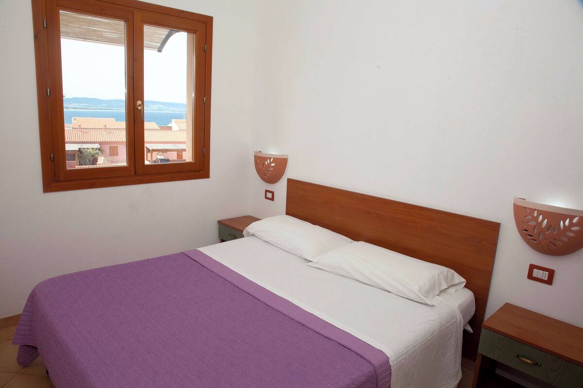 Une chambre à coucher comprenant un lit, une table de chevet, une lampe fixée au mur et une fenêtre donnant sur des immeubles et la mer.