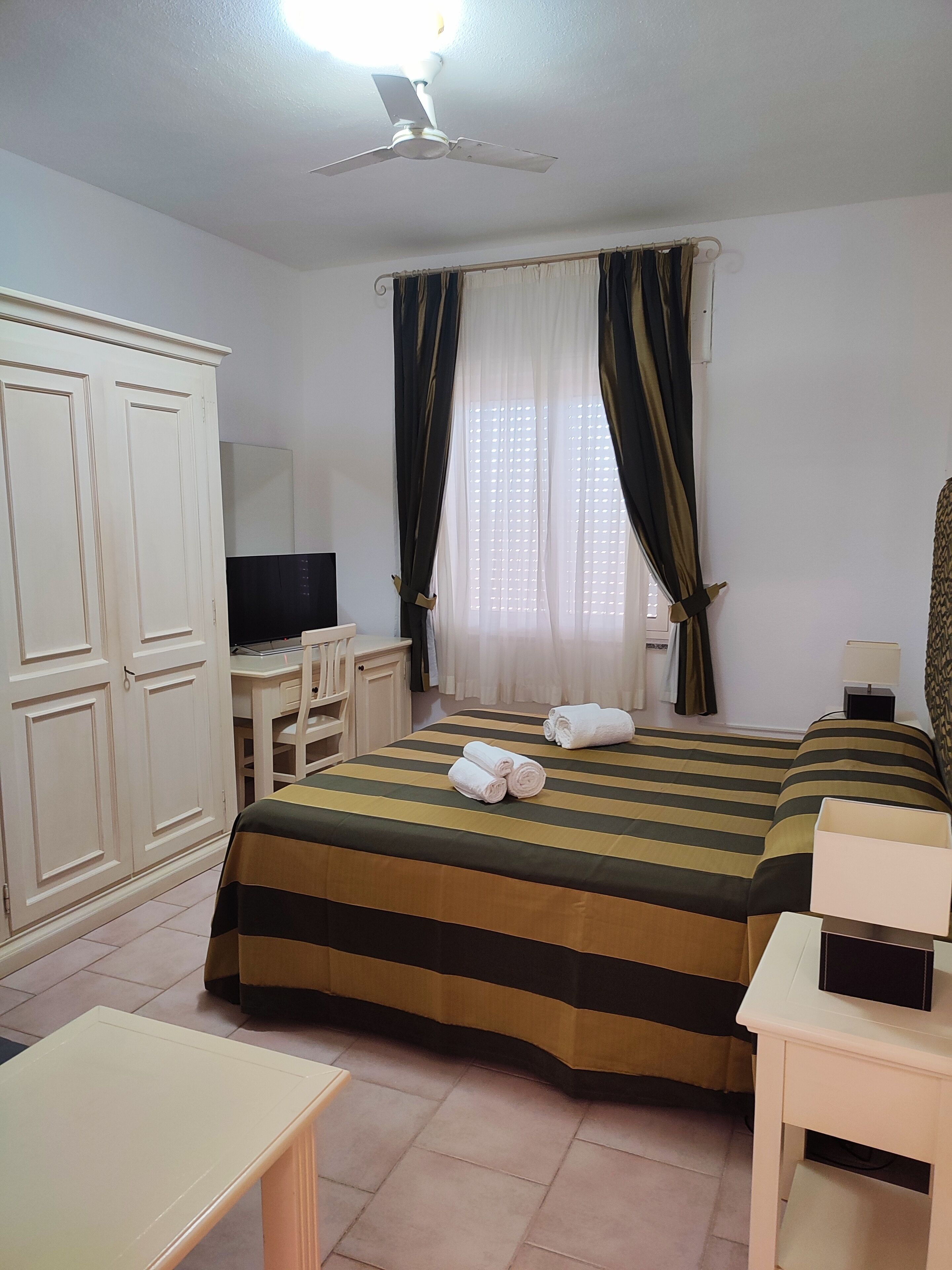 Ein Hotelzimmer mit Bett, Schreibtisch, Kleiderschrank, Deckenventilator und einem Fenster mit Vorhängen.