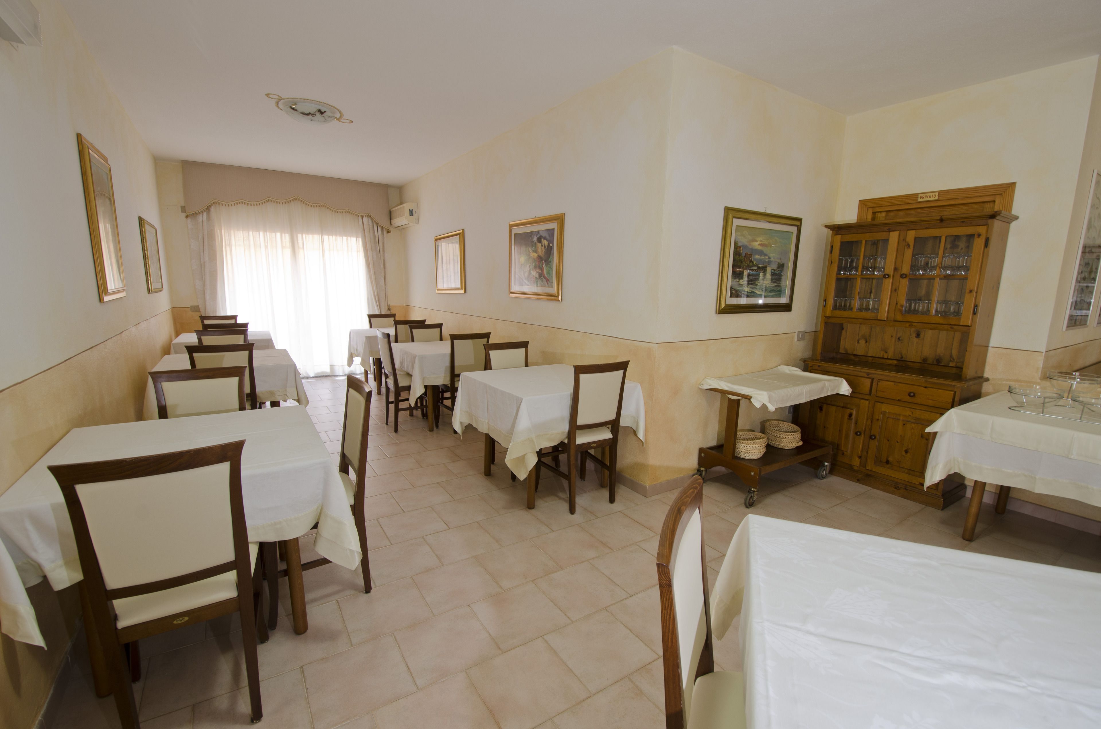 A dining area with multiple tables and chairs, a wooden cabinet with glassware, and framed artwork on the walls.