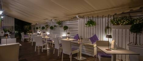 An outdoor dining area with white wicker furniture, tables, and chairs, under a white tent.