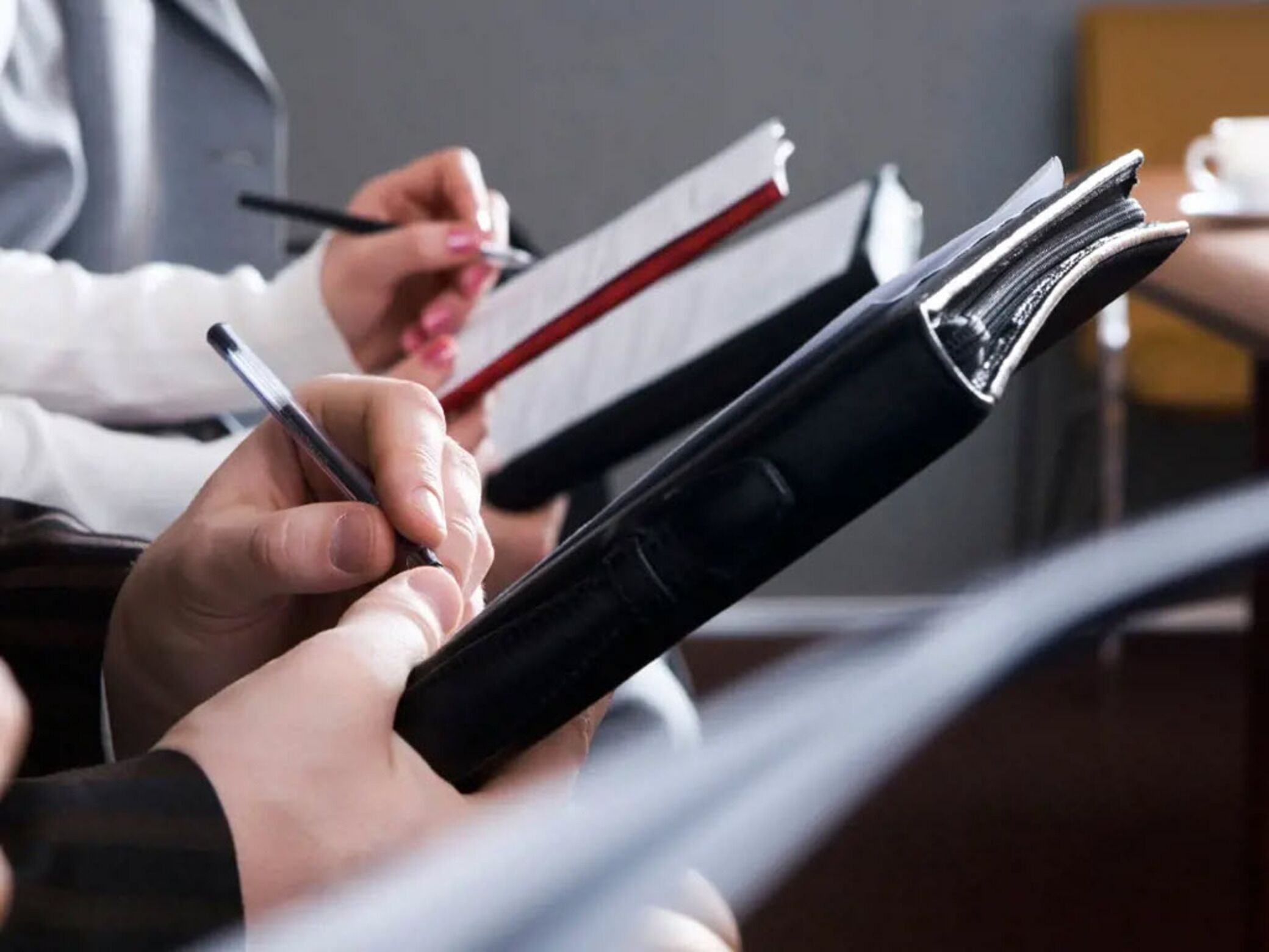 Een close-up van handen die een zwart notitieboekje en een pen vasthouden, terwijl een andere hand in een rood notitieboekje schrijft.