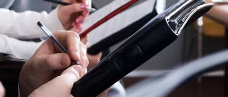 Een close-up van handen die een zwart notitieboekje en een pen vasthouden, terwijl een andere hand in een rood notitieboekje schrijft.