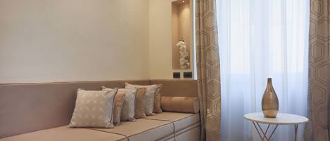 A modern living room with a beige sofa, patterned cushions, a small white table with a vase, and sheer curtains.