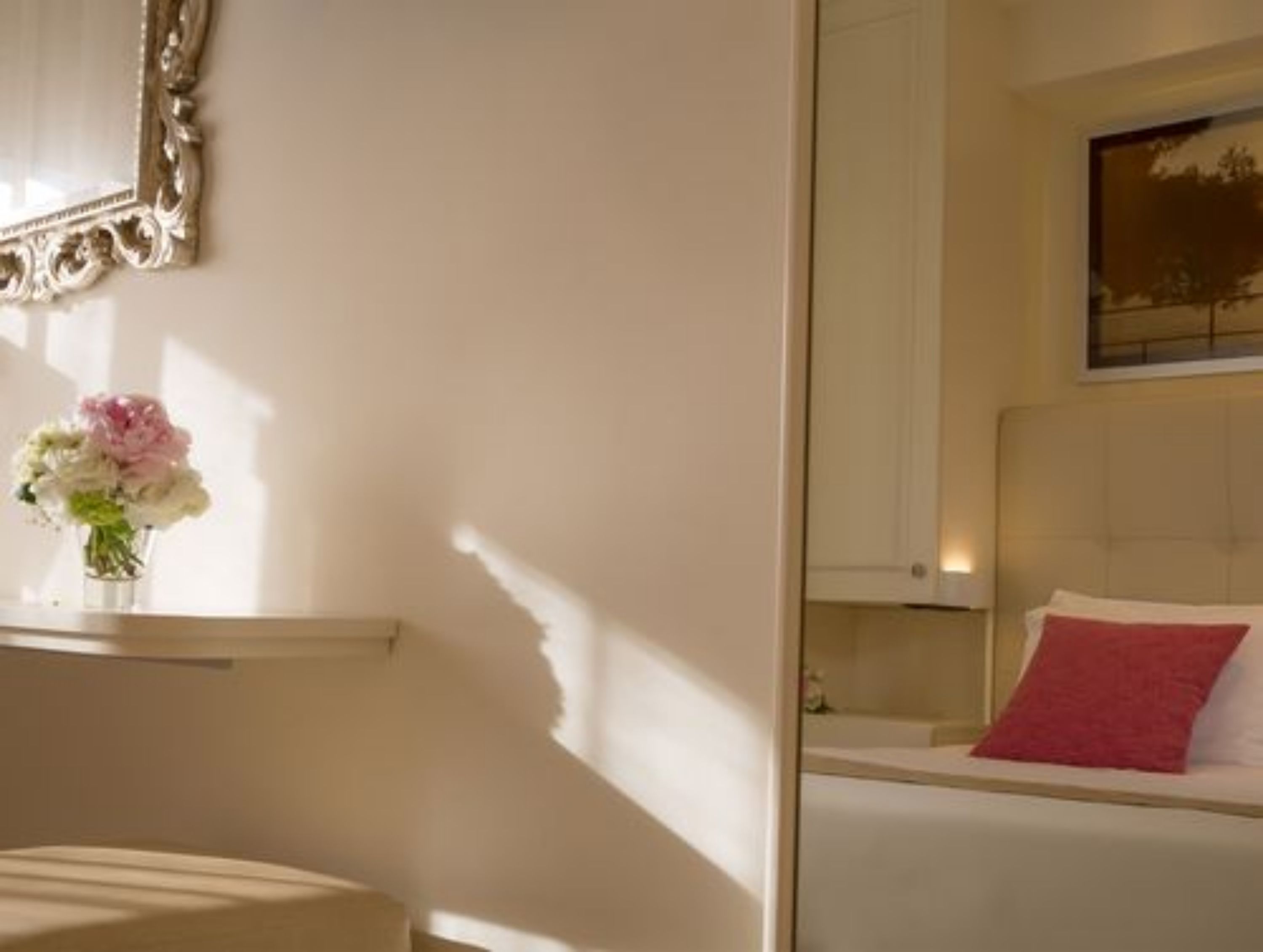 Een hotelkamer met een bed, nachtkastje, vaas met bloemen en een spiegel met een sierlijk frame.