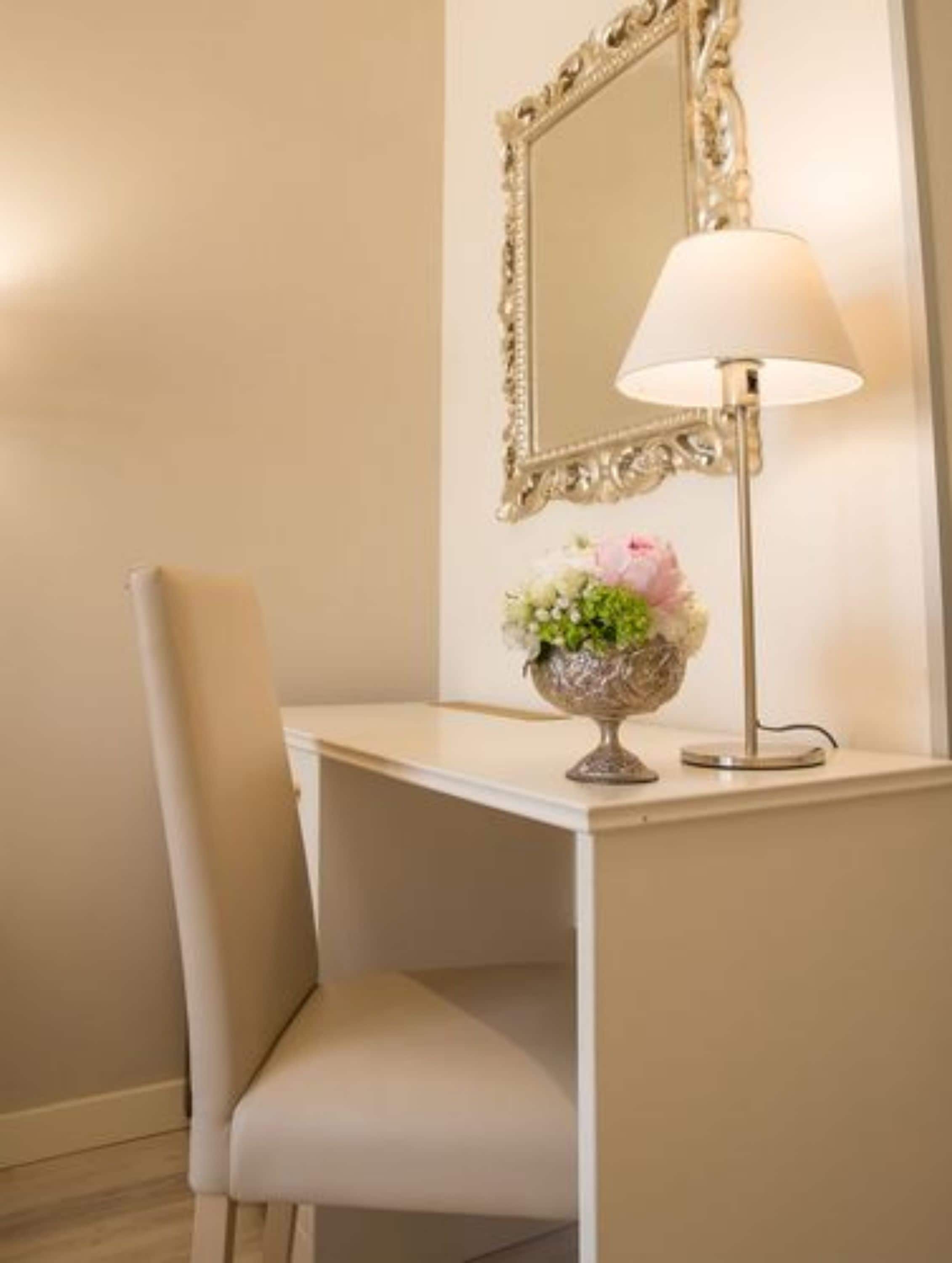Een hoek van een kamer met een wit bureau, een stoel, een lamp en een spiegel met een versierd frame.