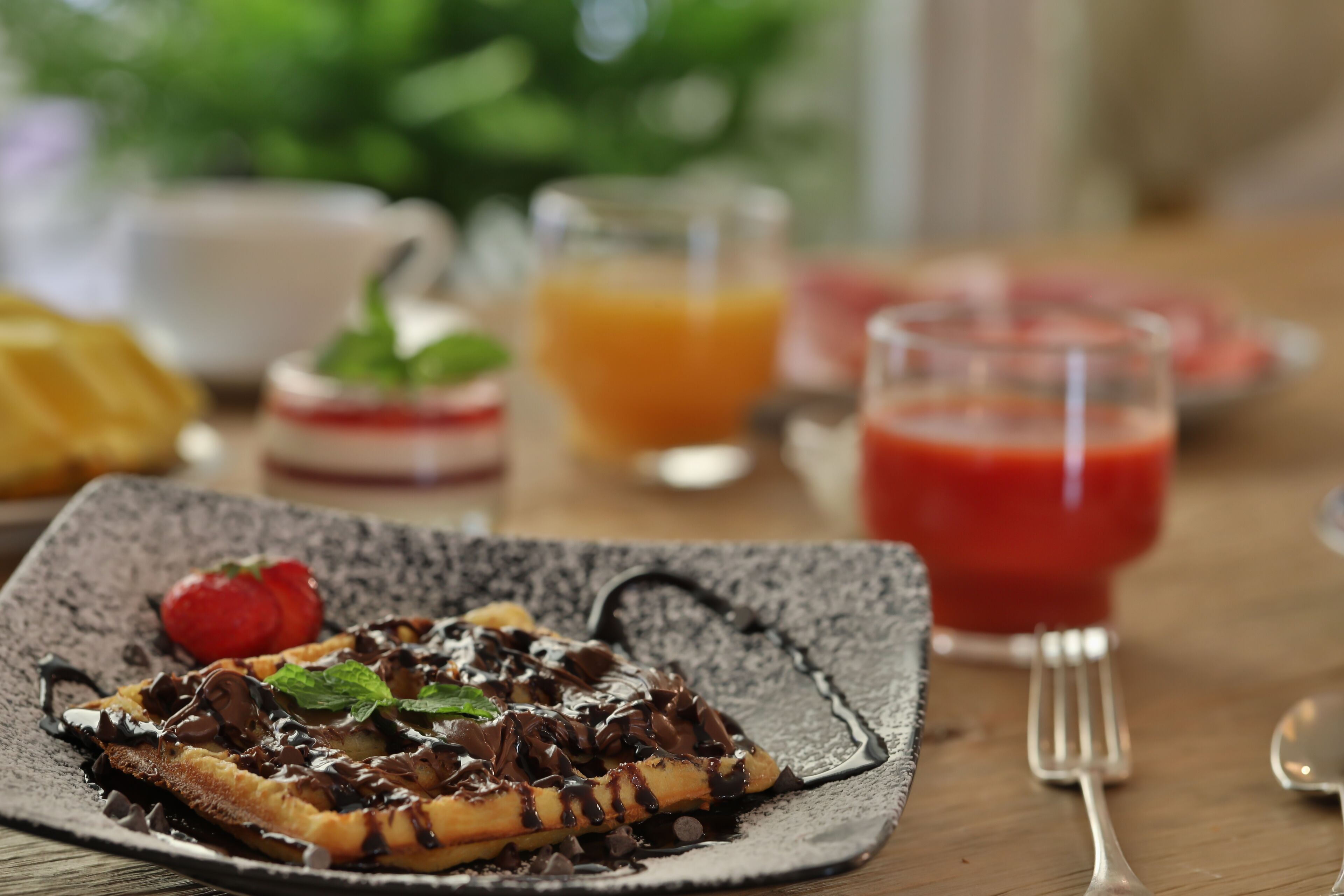 A breakfast setting with a plate of waffles topped with chocolate and a strawberry, accompanied by a glass of orange juice and a glass of red juice.