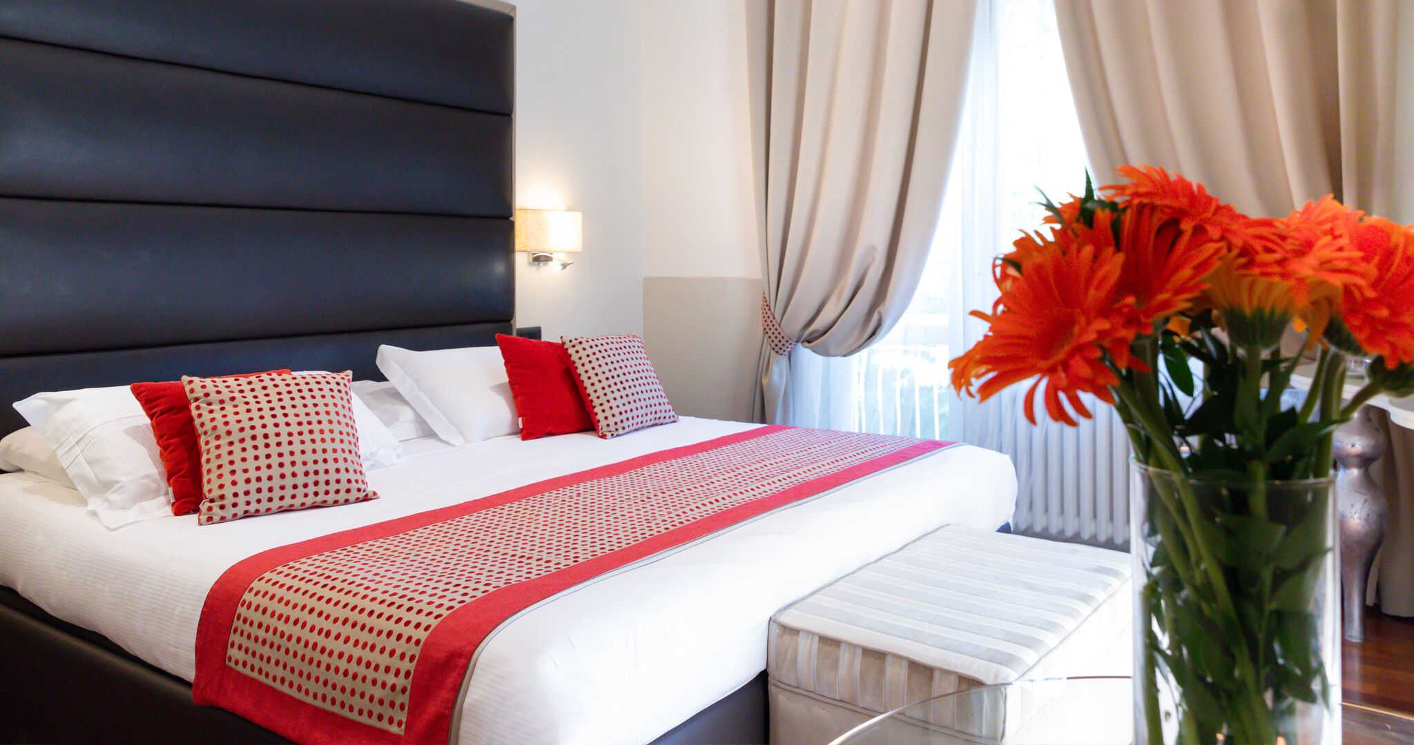 Habitación de hotel con una cama grande, una mesita de noche, un florero con flores y una ventana con cortinas.