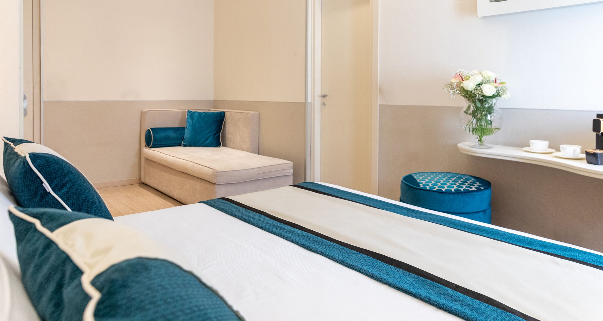 Een moderne hotelkamer met een bed, een klein zithoekje met een kussen, een nachtkastje met een bloemenvaas en een koffiezetapparaat.
