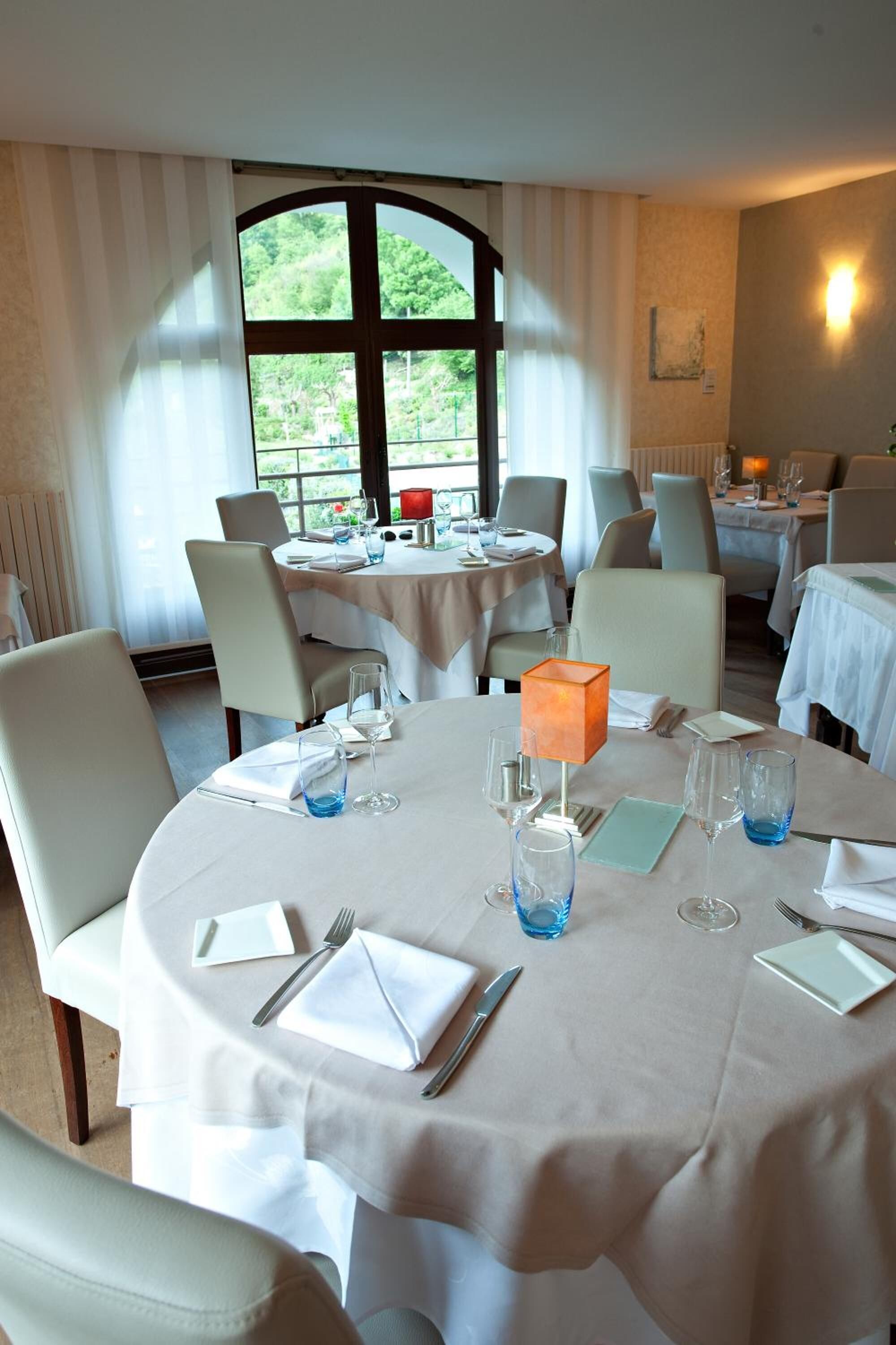 Un espace repas avec des tables rondes, des chaises blanches et une grande fenêtre donnant sur un paysage verdoyant.