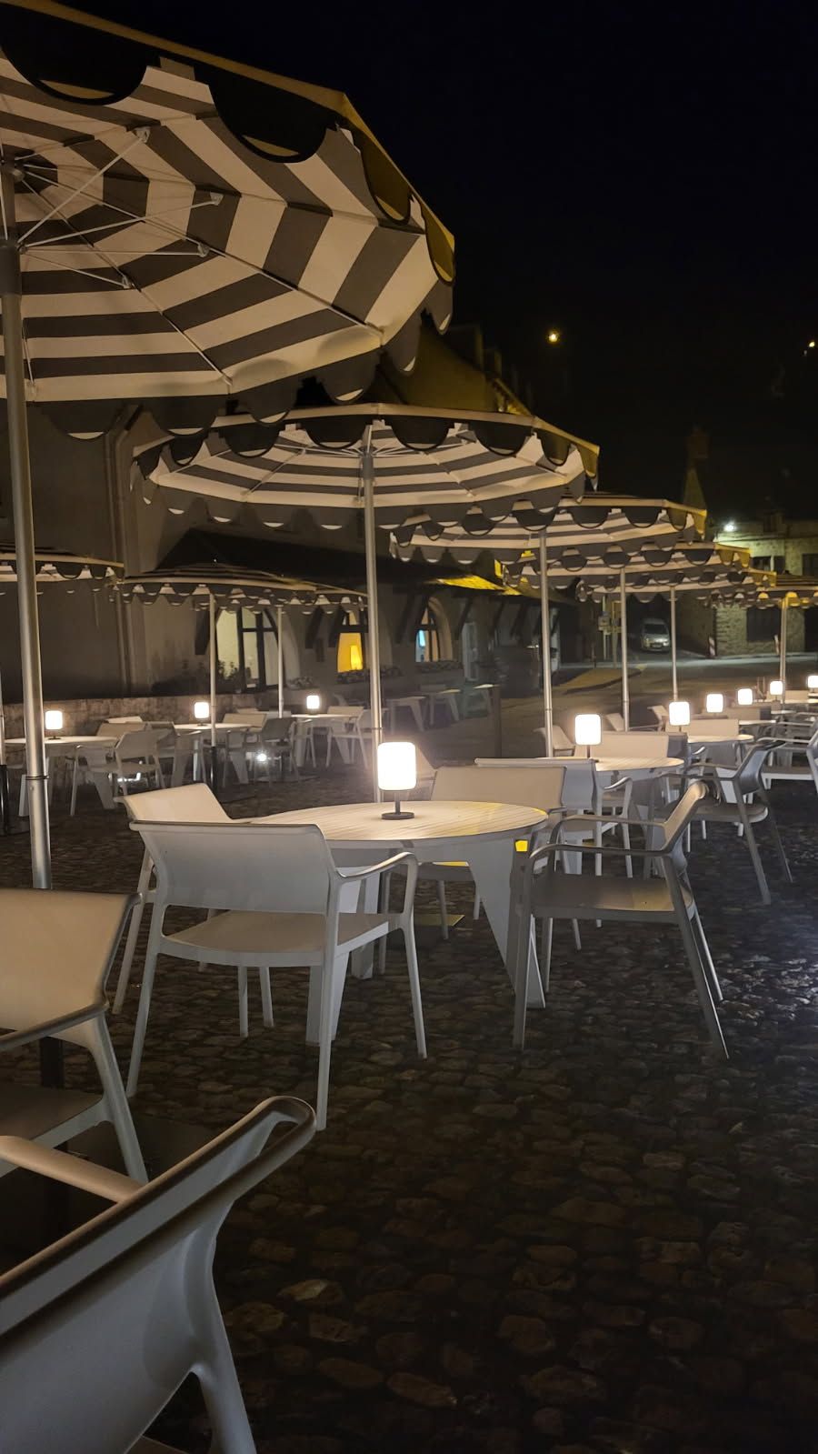 Un área de comedor al aire libre con mesas y sillas bajo sombrillas a rayas, sobre un suelo adoquinado.