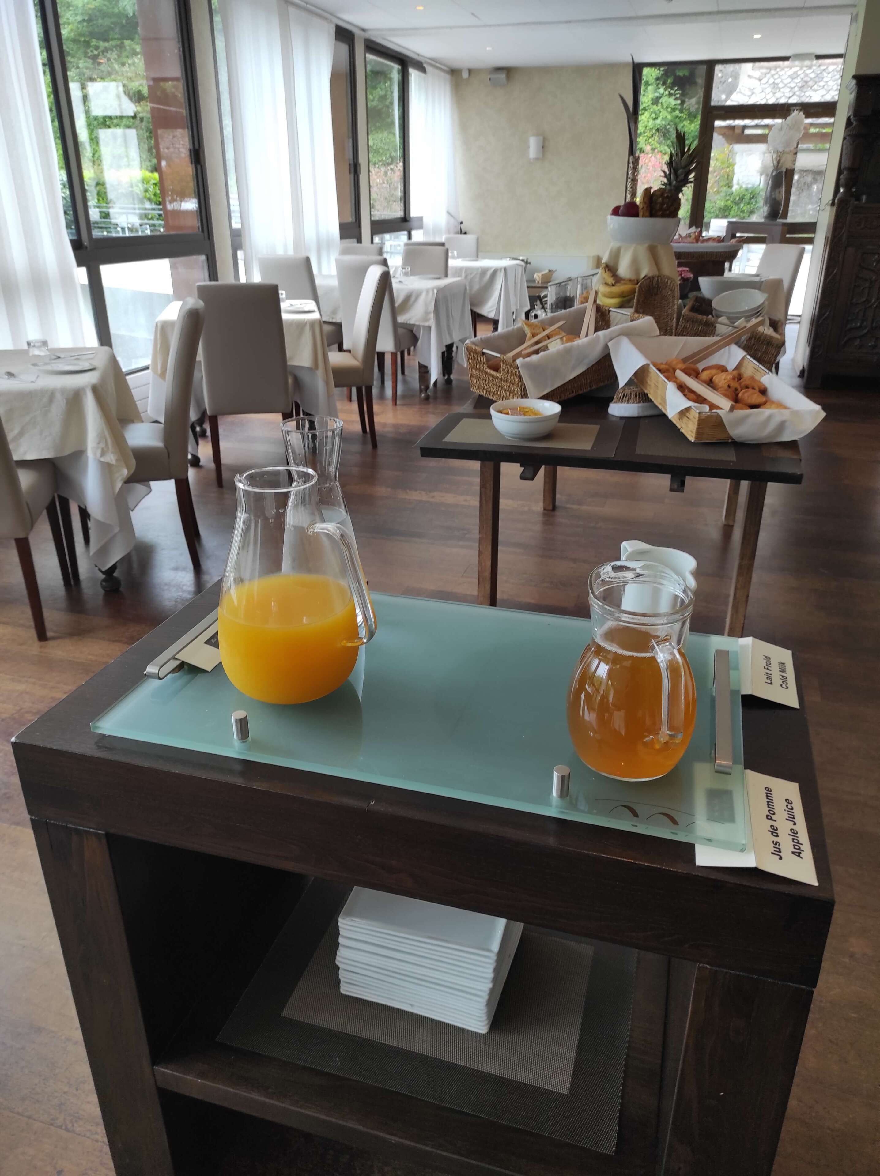Une table mise pour le petit-déjeuner, avec un panier de fruits, un panier de pain et une carafe de jus.
