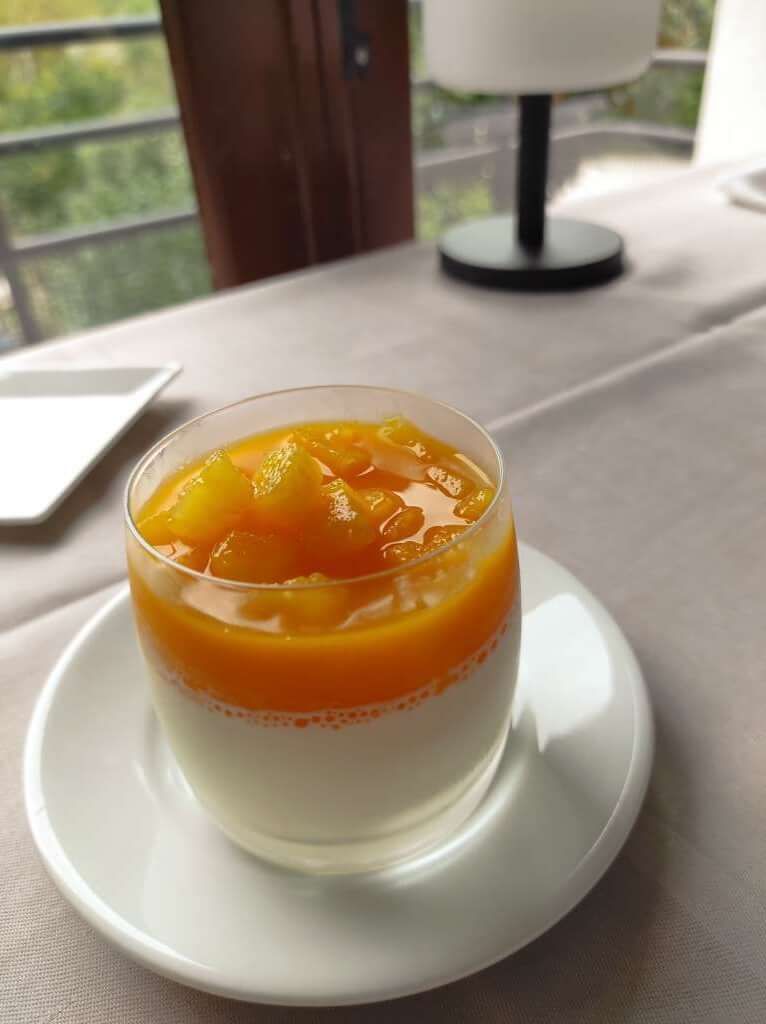 A dessert in a glass on a white plate, with a lamp and a window in the background.