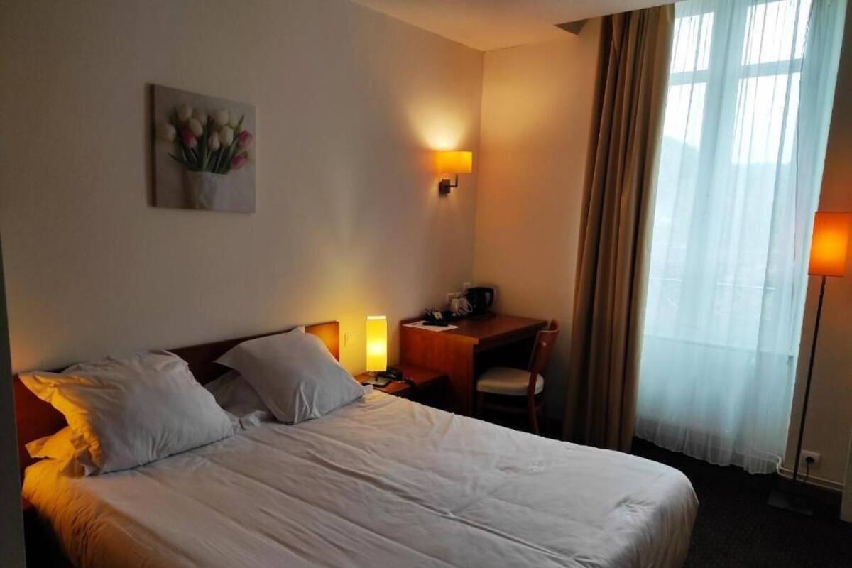 Una habitación de hotel con una cama, lámparas de noche, un escritorio con una silla, un cuadro en la pared y una ventana con cortinas.