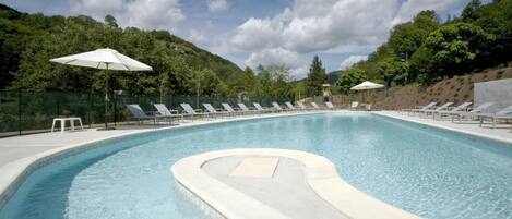 A swimming pool with lounge chairs and umbrellas, surrounded by greenery and hills.
