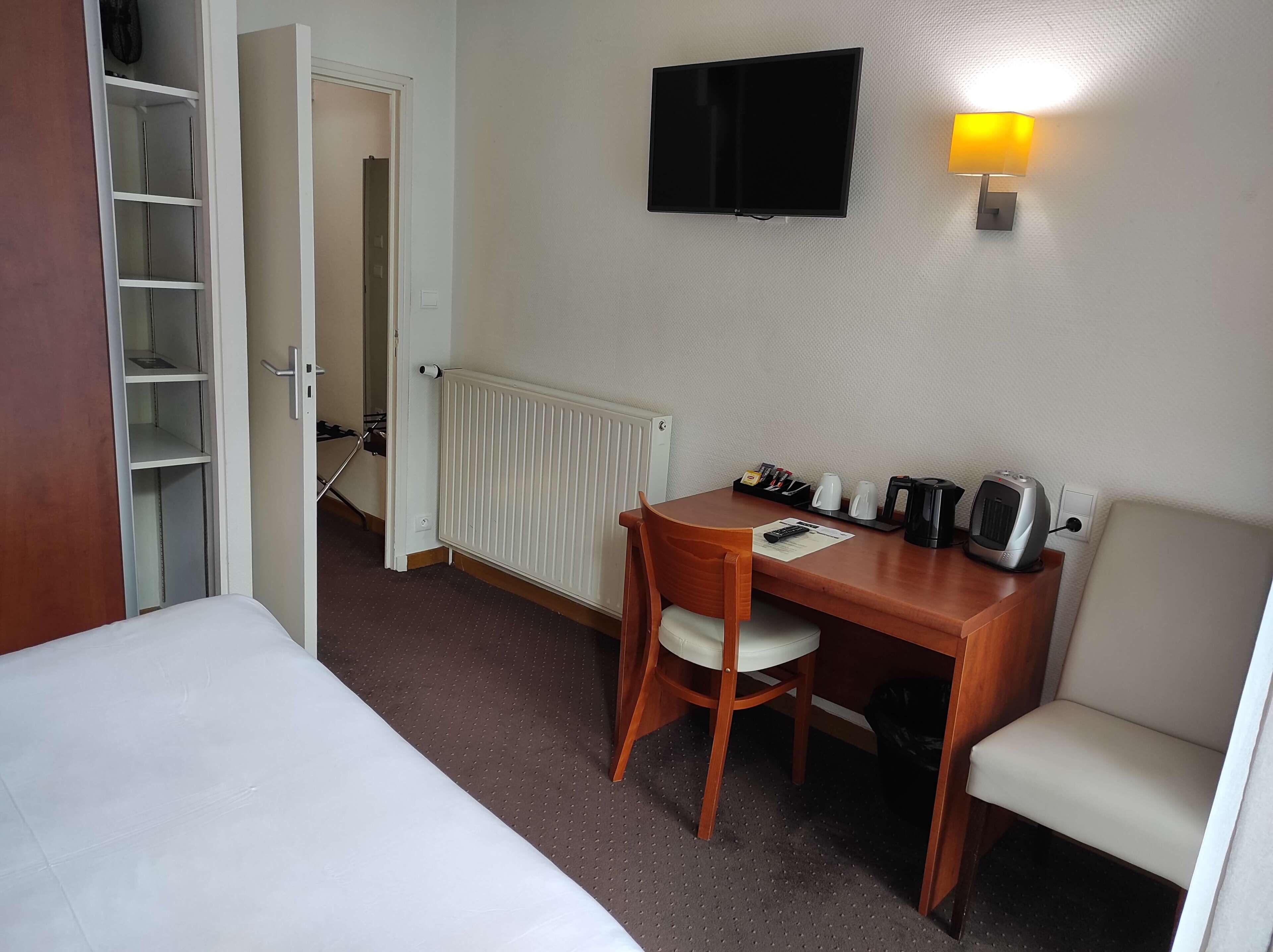 A hotel room with a bed, a desk with a chair, a television, a wall-mounted lamp, and a radiator.
