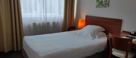 Quarto de hotel com cama, escrivaninha, cadeira, luminária e um quadro na parede.