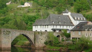 Eine Steinbrücke über einem ruhigen Fluss, umgeben von üppiger Vegetation und einem Hügel mit Gebäuden.
