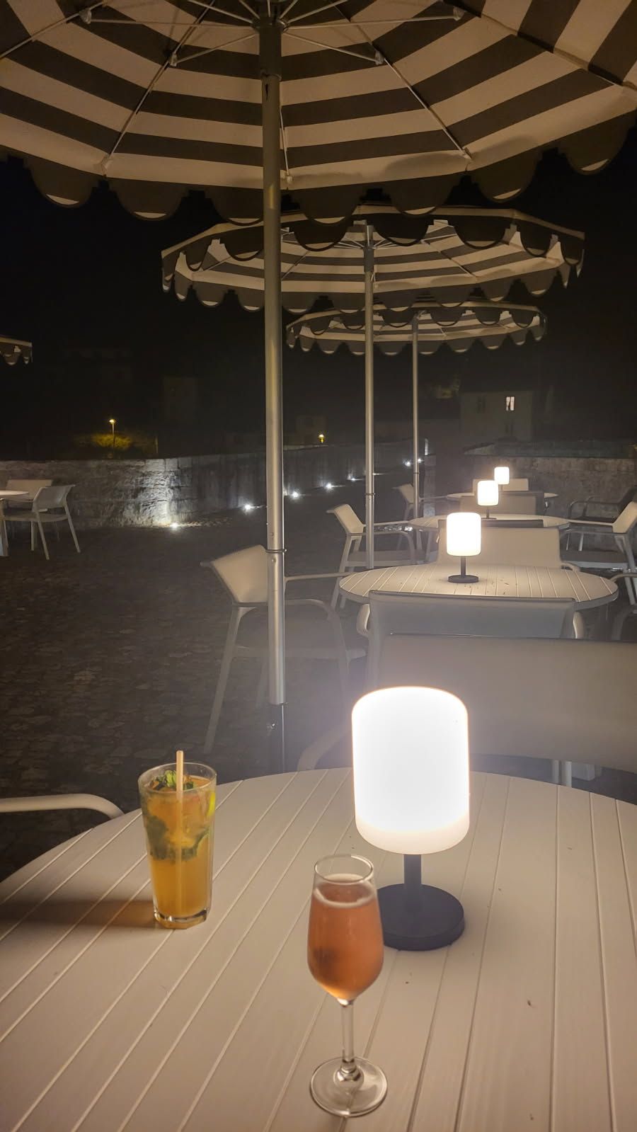 Ein Sitzbereich im Freien mit Tischen und Stühlen unter gestreiften Sonnenschirmen, ein Glas Eistee und ein Weinglas auf einem Tisch.
