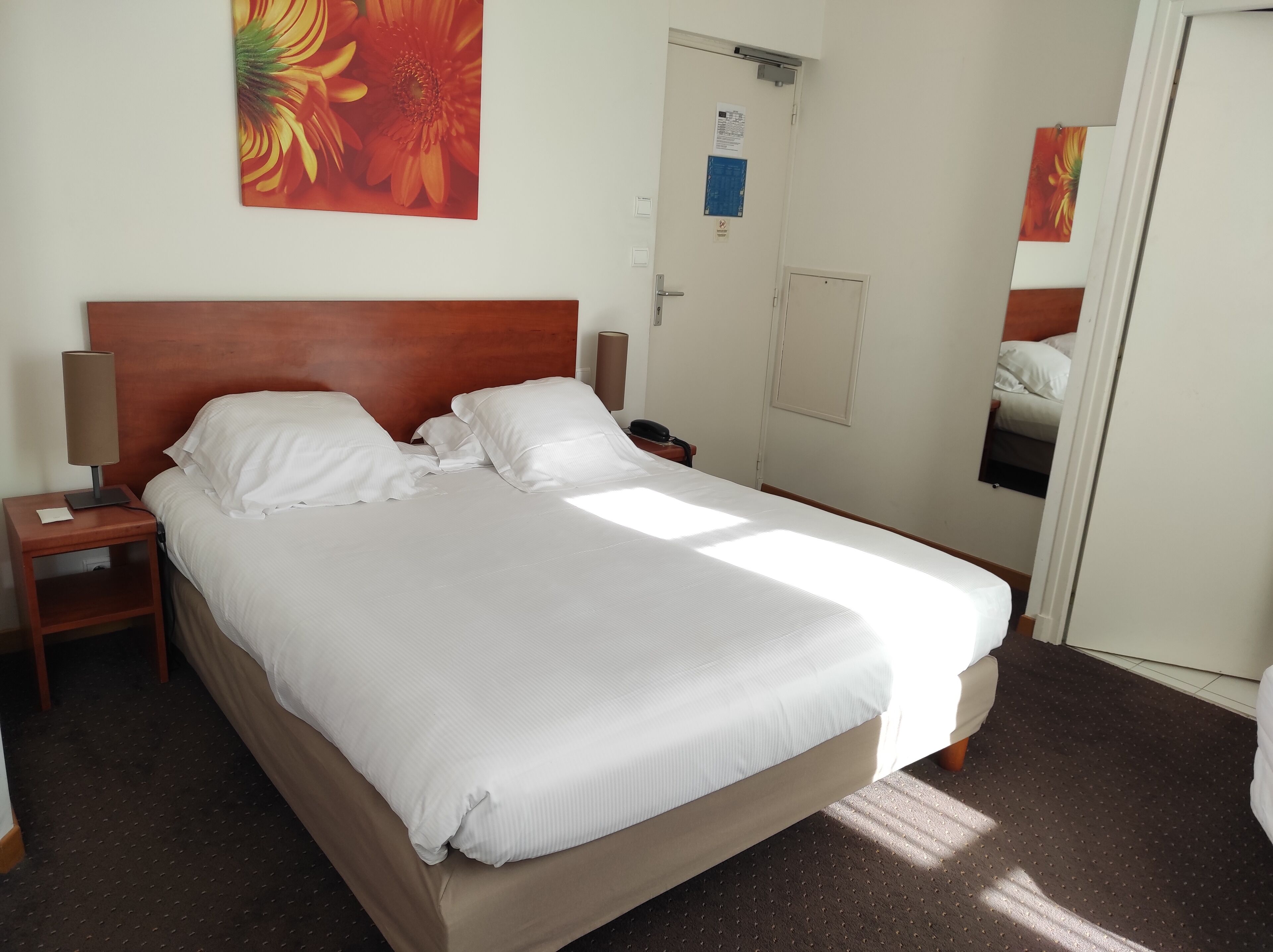 A neatly made bed with white linens, a wooden headboard, a nightstand with a lamp, and a wall-mounted mirror.