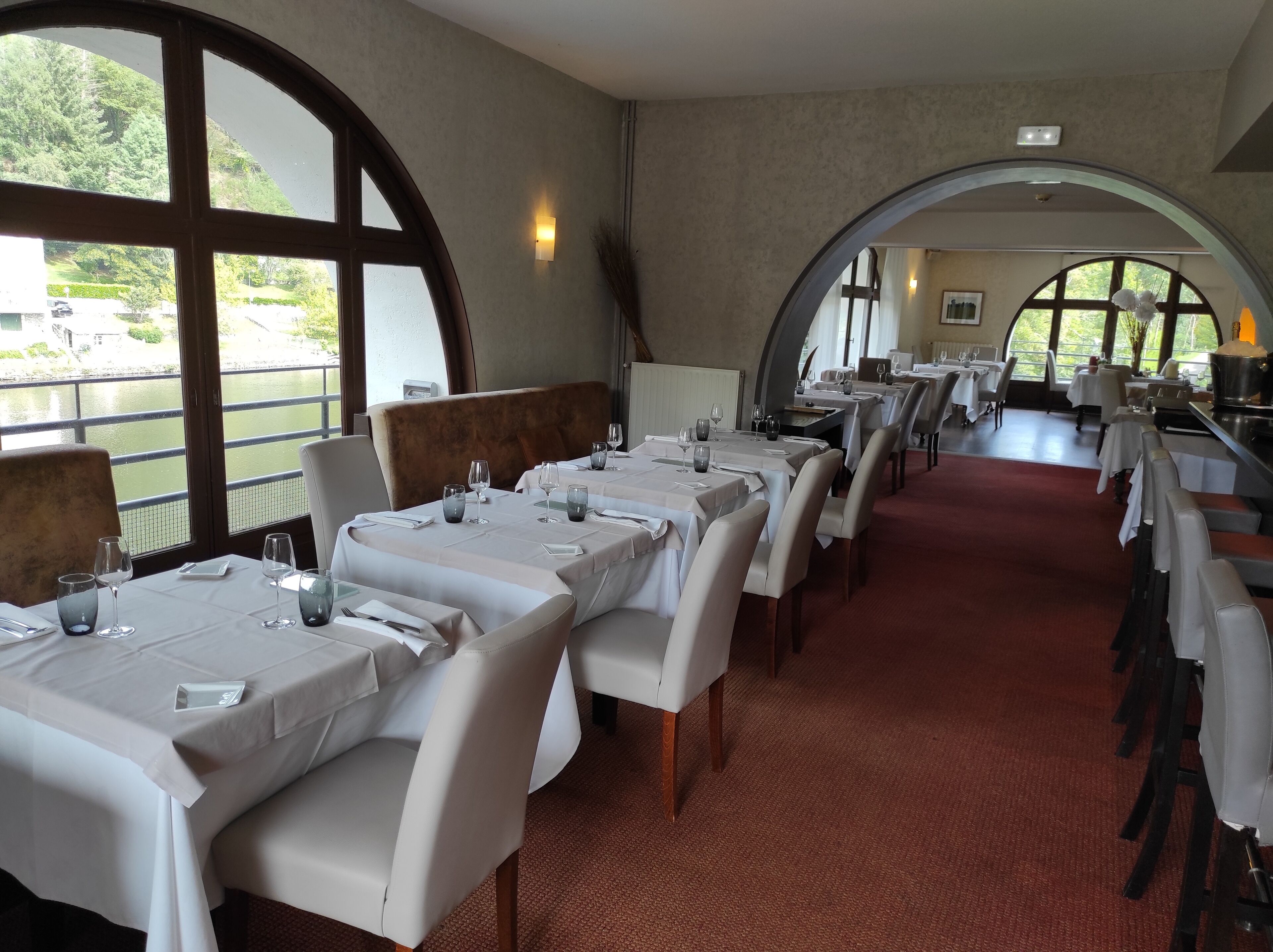 Een restaurant met meerdere tafels, witte tafelkleden en stoelen. Grote ramen bieden uitzicht op groen en een waterpartij.