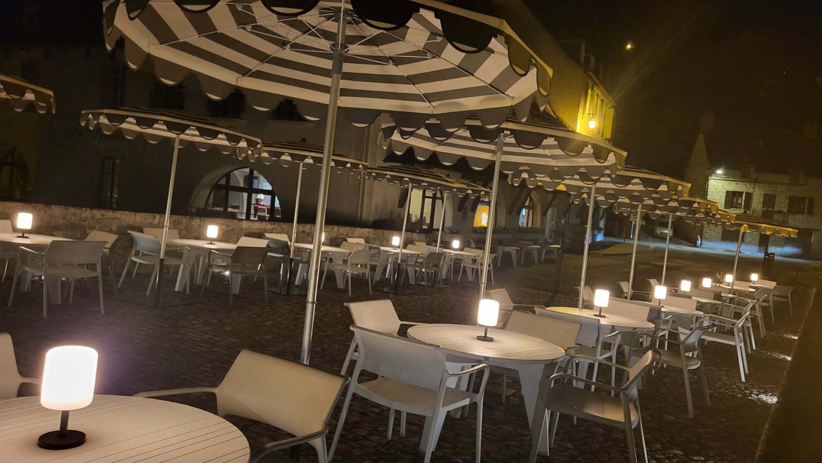An outdoor dining area with round tables and chairs, covered by striped umbrellas, and illuminated by table lamps.