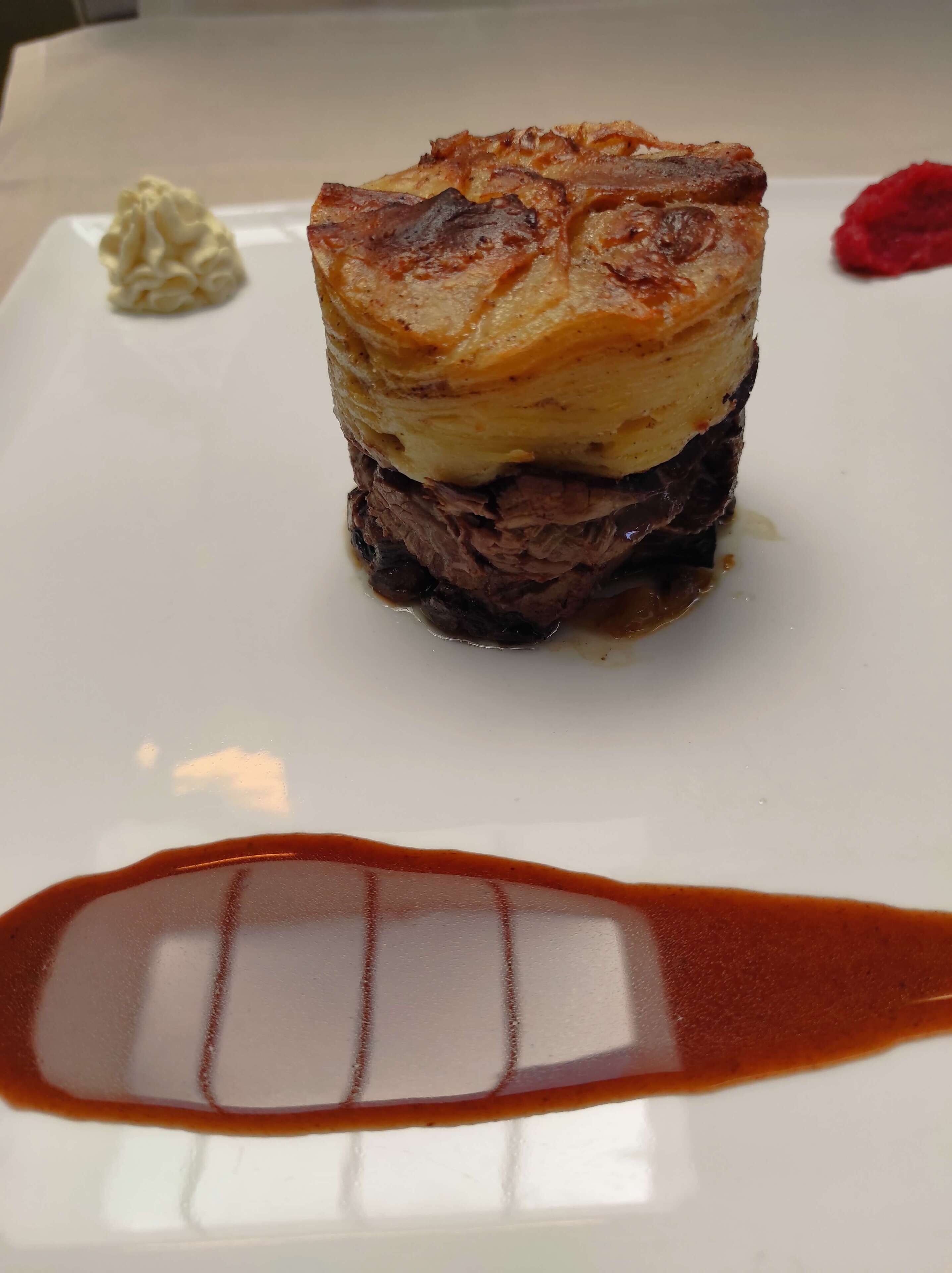 A dish with a golden-brown crust, a layer of mashed potatoes, and a dark meat base, served on a white plate with a red sauce.