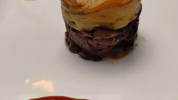 A dish with a golden-brown crust, a layer of mashed potatoes, and a dark meat base, served on a white plate with a red sauce.