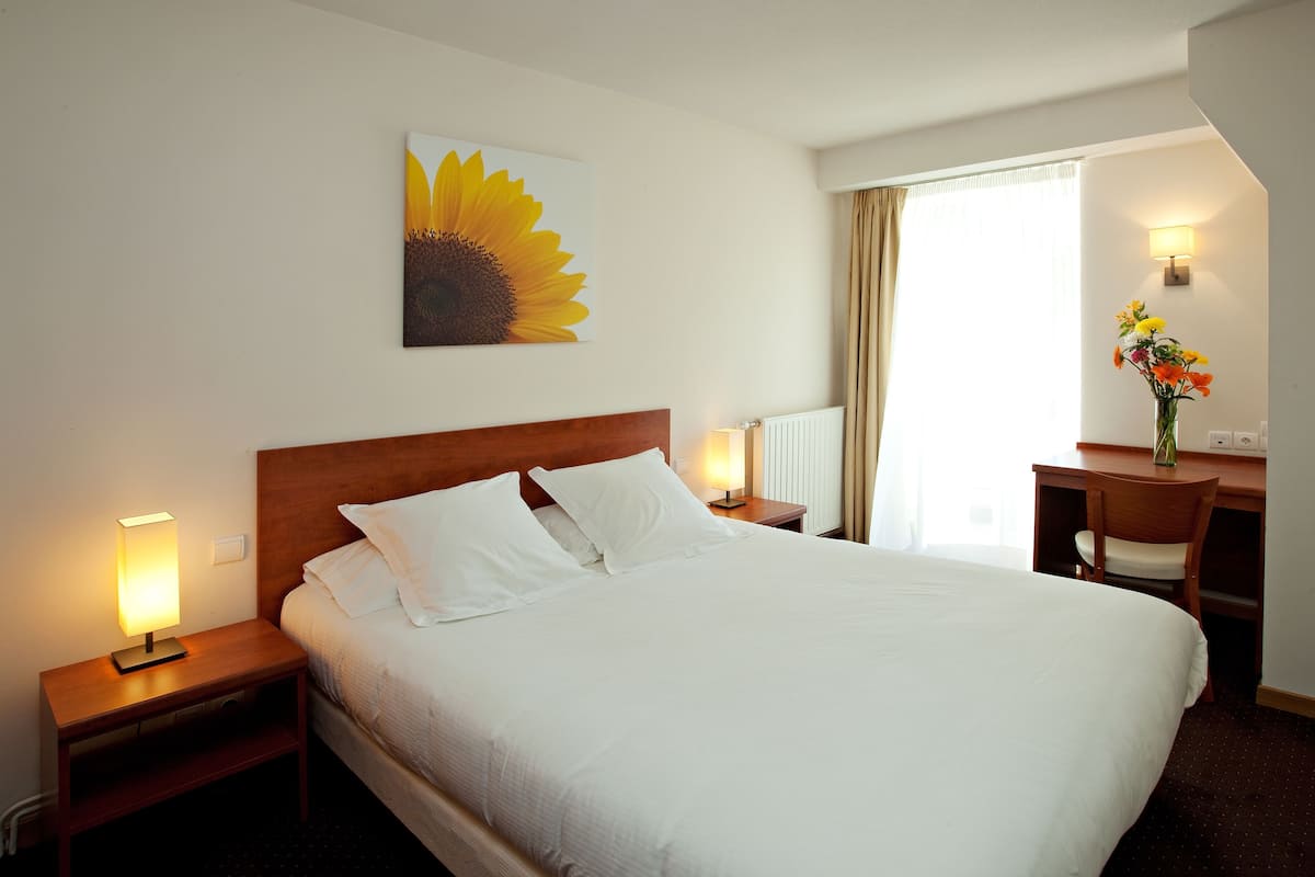 Habitación de hotel con una cama grande, un escritorio con una silla, una lámpara, una obra de arte colgando en la pared que representa un girasol, y una ventana con cortinas.
