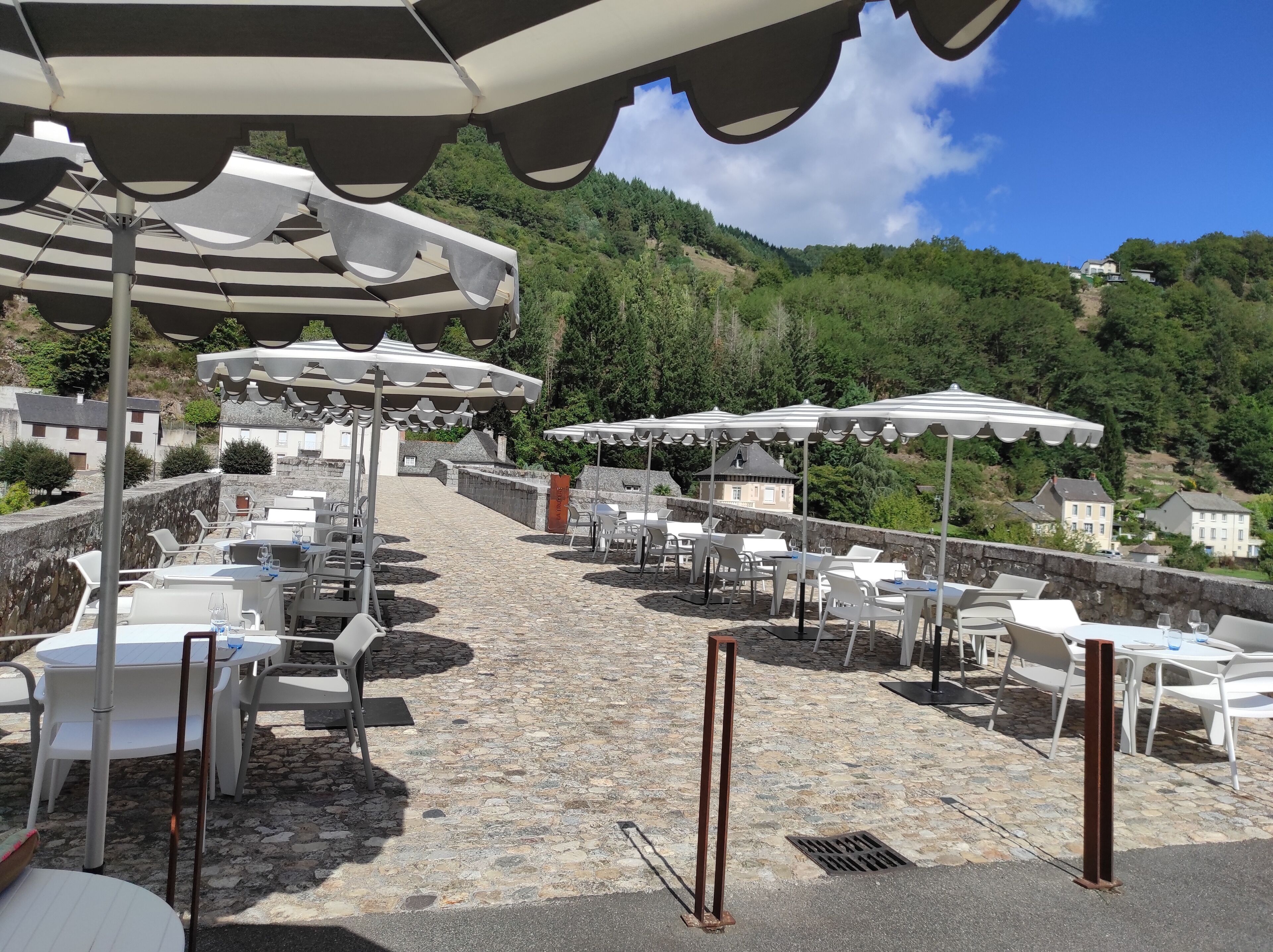 Een terras buiten met stenen vloer, witte tafels en stoelen, en gestreepte parasols.