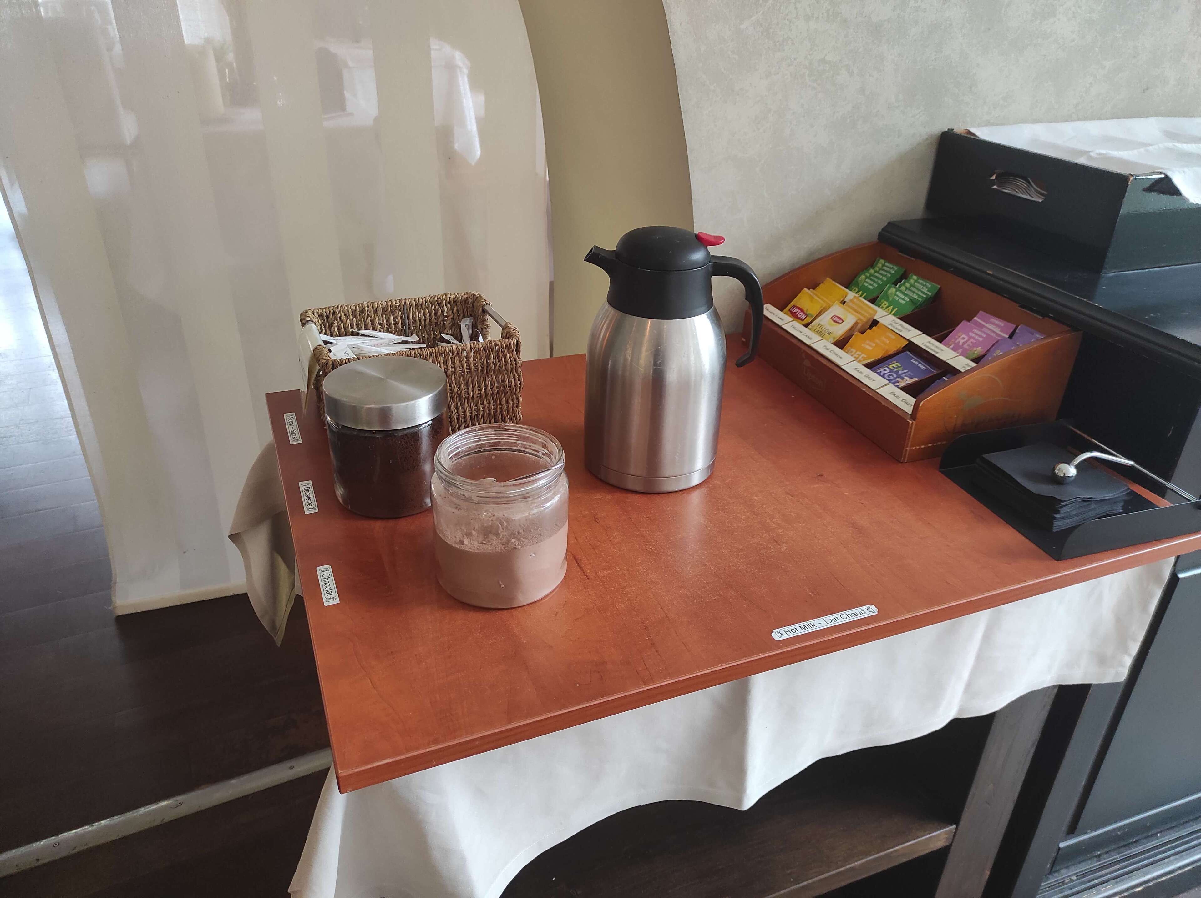 A coffee station with a coffee maker, a thermos, and a jar of hot chocolate on a wooden table.