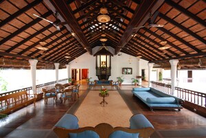 Un hall espacioso con suelo de madera, un techo alto con vigas a la vista, una zona de estar central con sofás y sillones, y una lámpara decorativa.