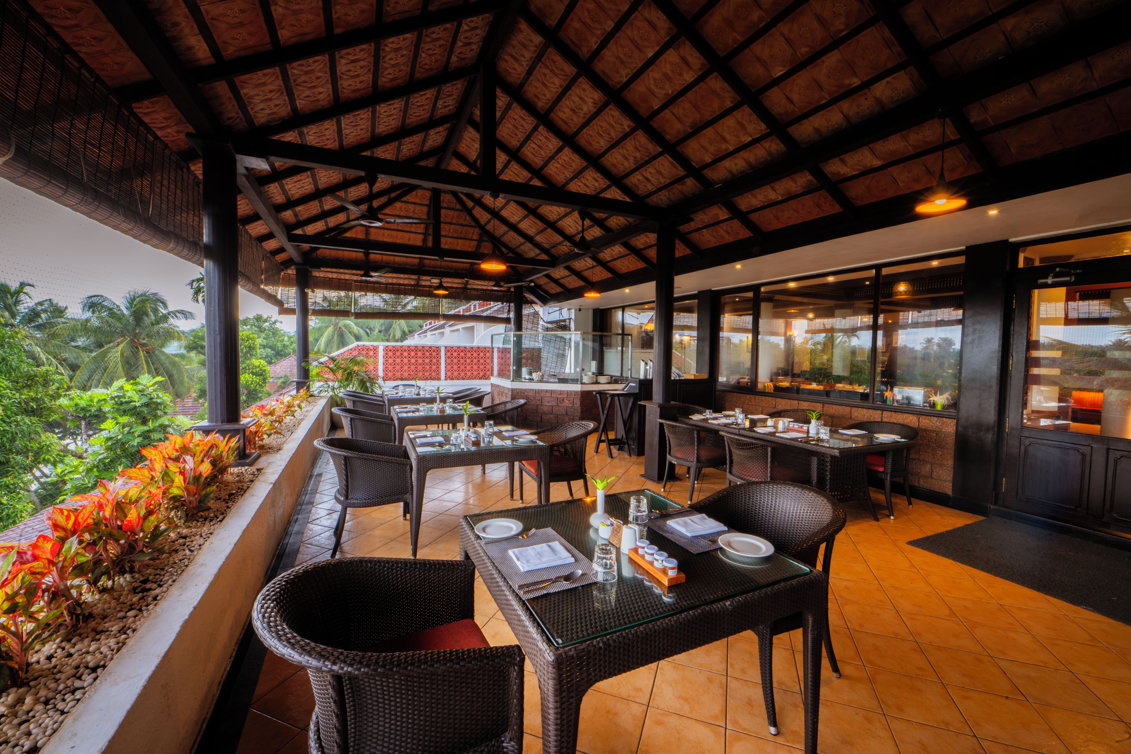 An outdoor restaurant with tables and chairs, a covered area, and a view of palm trees and buildings in the background.