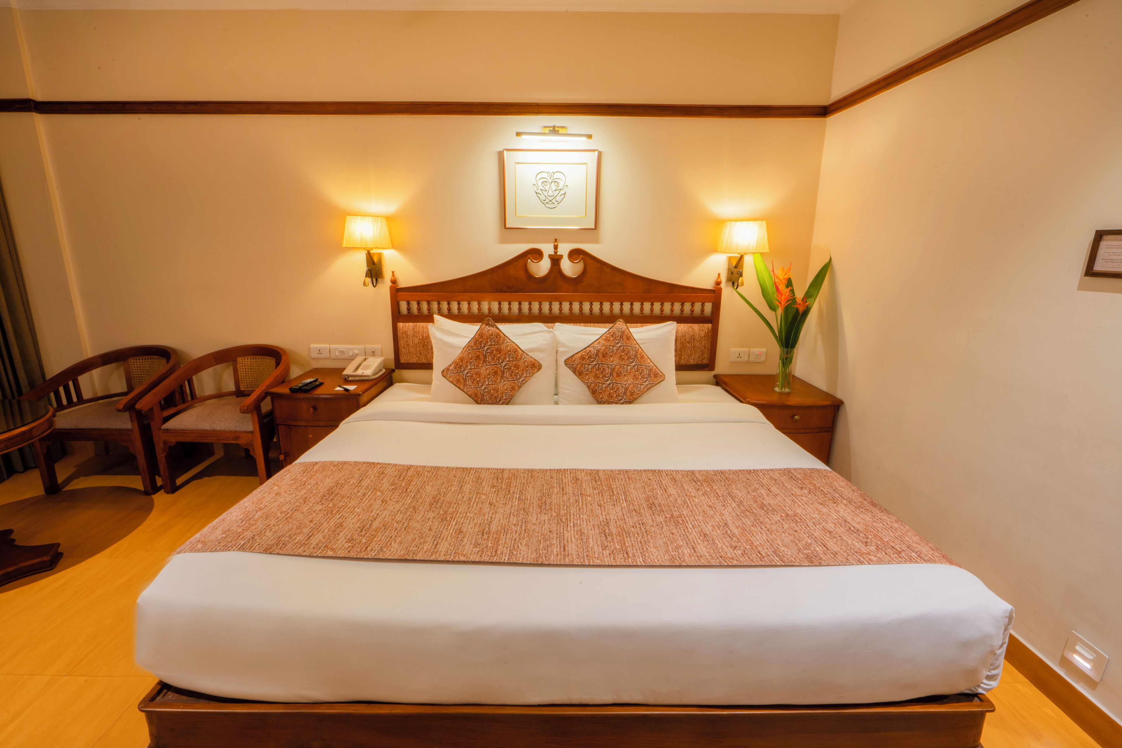 Habitación de hotel con una cama grande, cabecera de madera, dos lámparas de noche, una mesita de noche, una silla y un jarrón con flores.