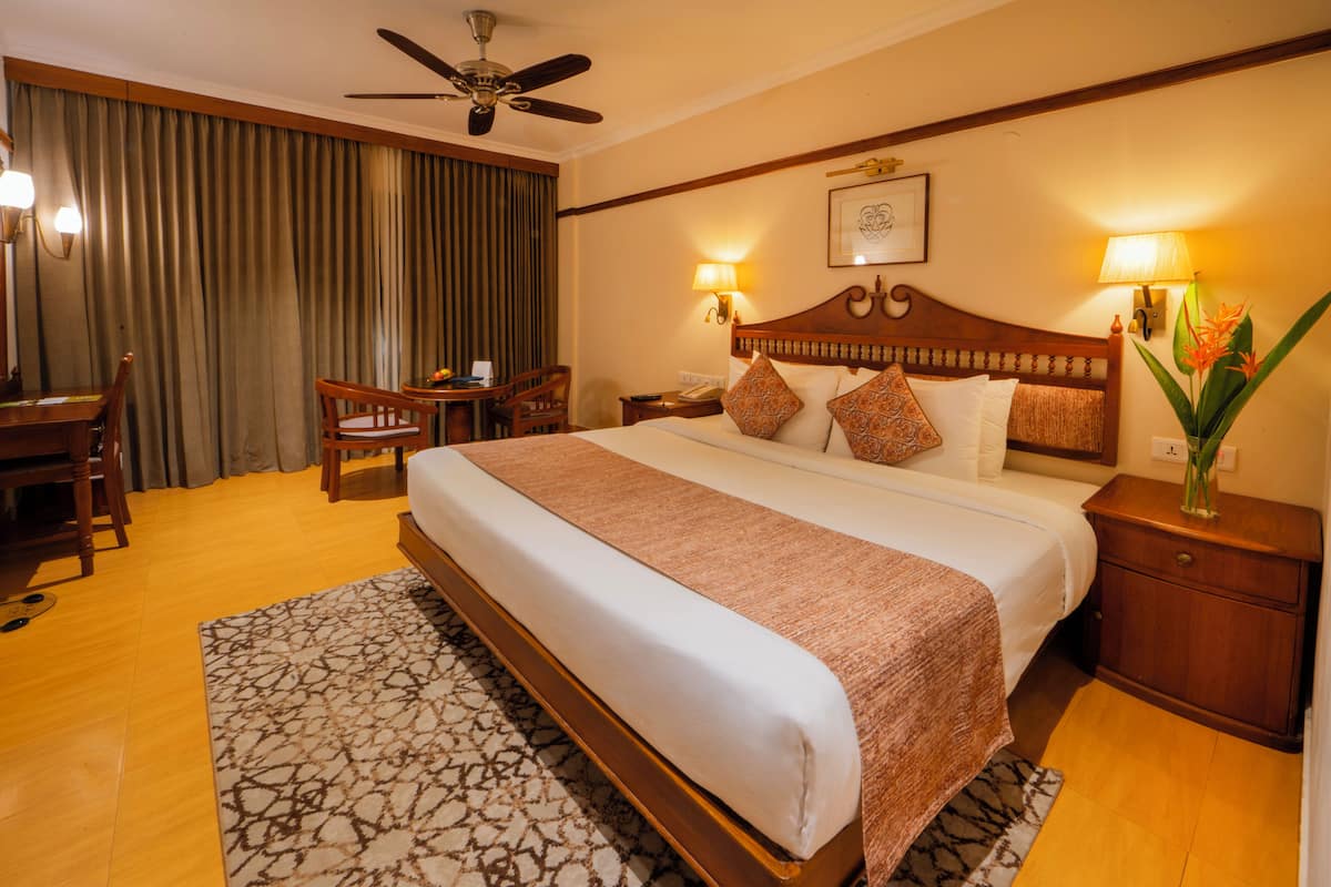 Ein Hotelzimmer mit einem großen Bett, einem Schreibtisch mit Stuhl, einem Nachttisch, einem Deckenventilator und einem großen Fenster mit Vorhängen.