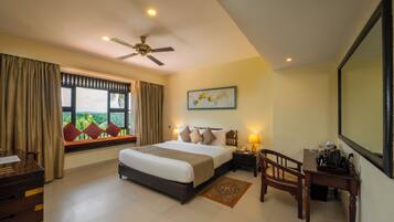 A hotel room with a large bed, a desk, a chair, a nightstand, a ceiling fan, a window with curtains, and a painting on the wall.